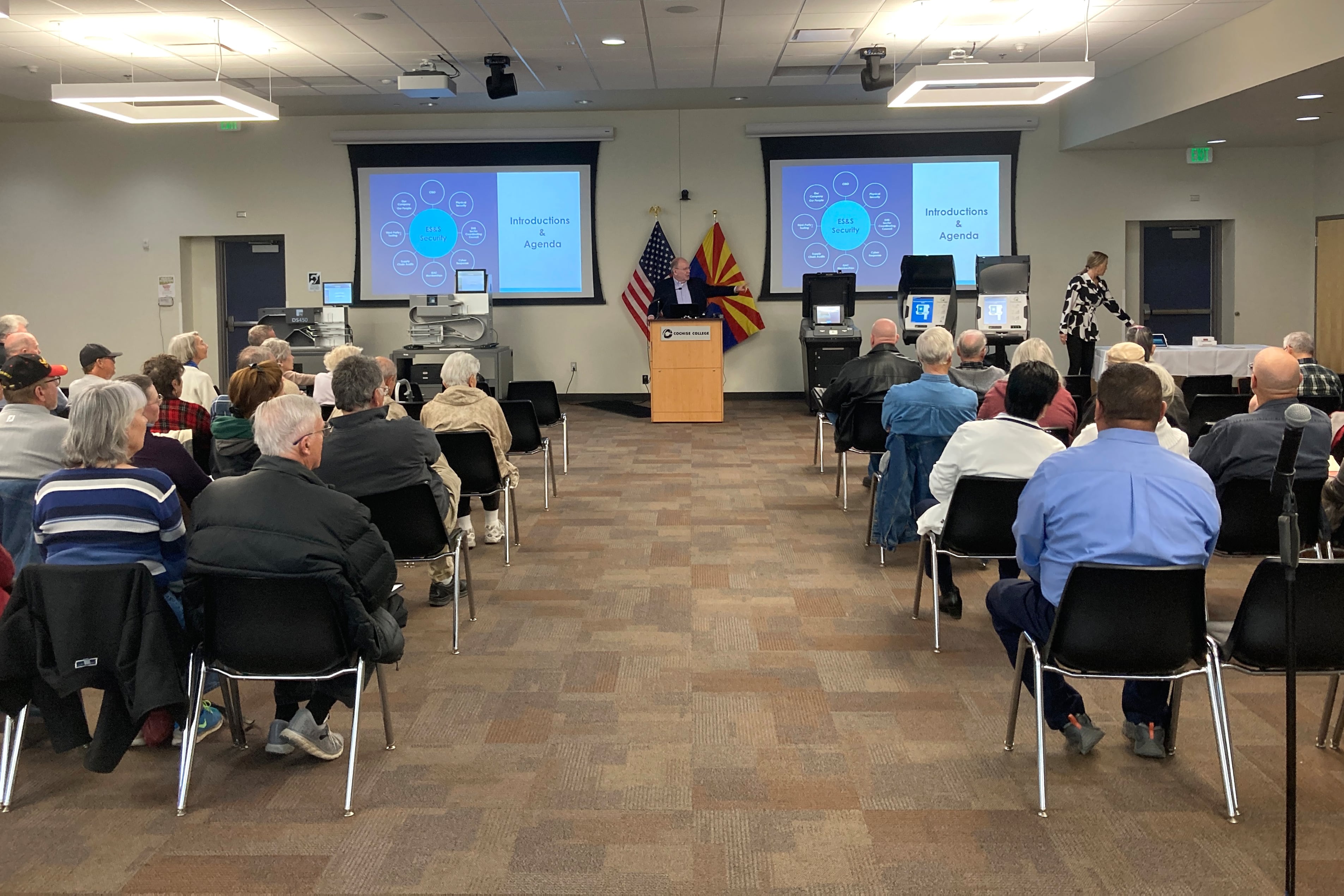 A crowd of people sit in chairs and look up at two screens.