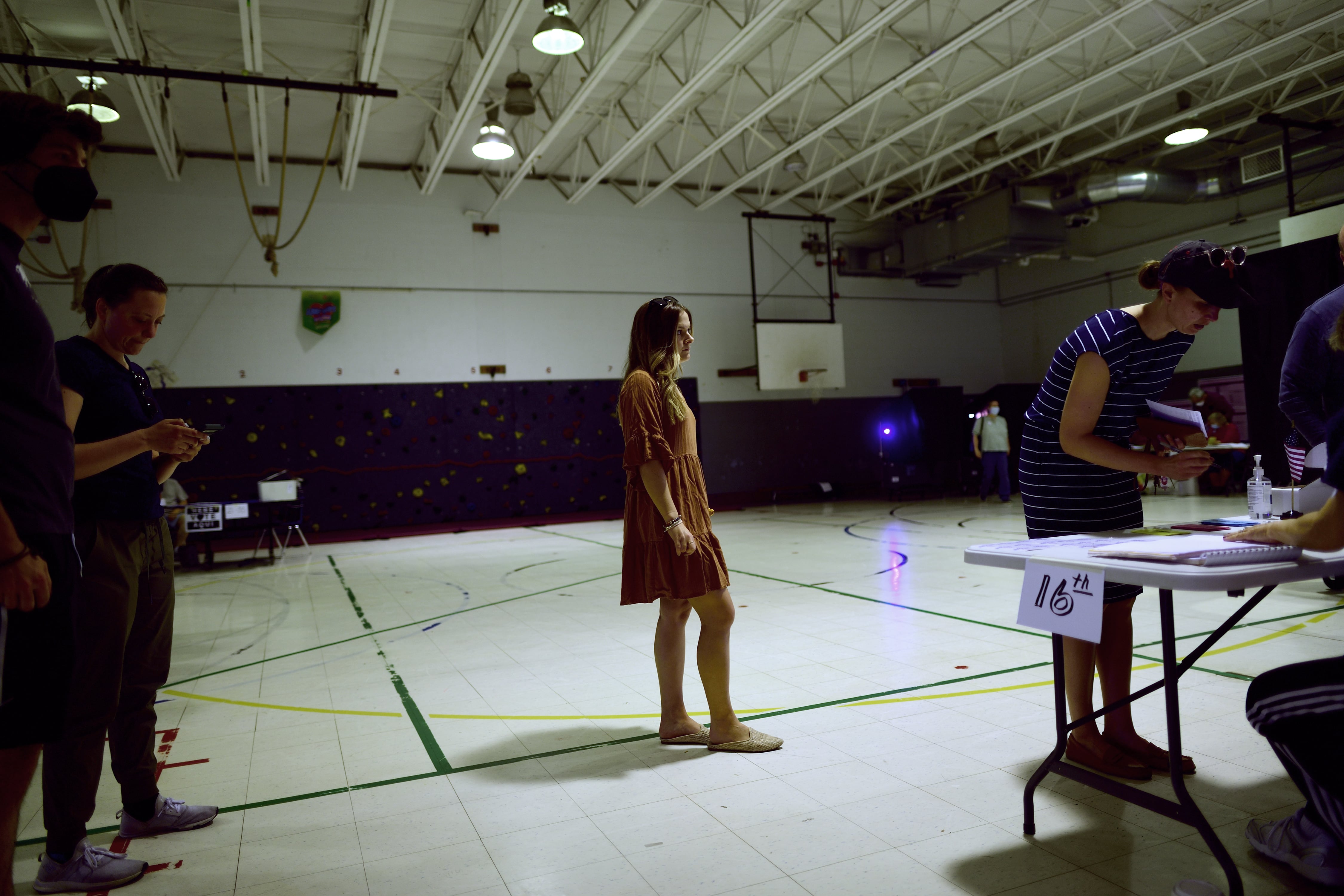 A woman with long dark hair waits before a table in a school gymnasium.