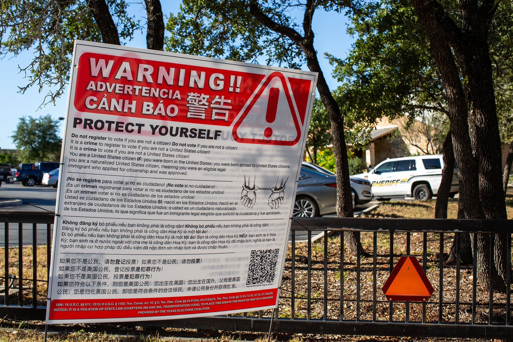 A large red and white warning sign in front of a fence and parked cars.