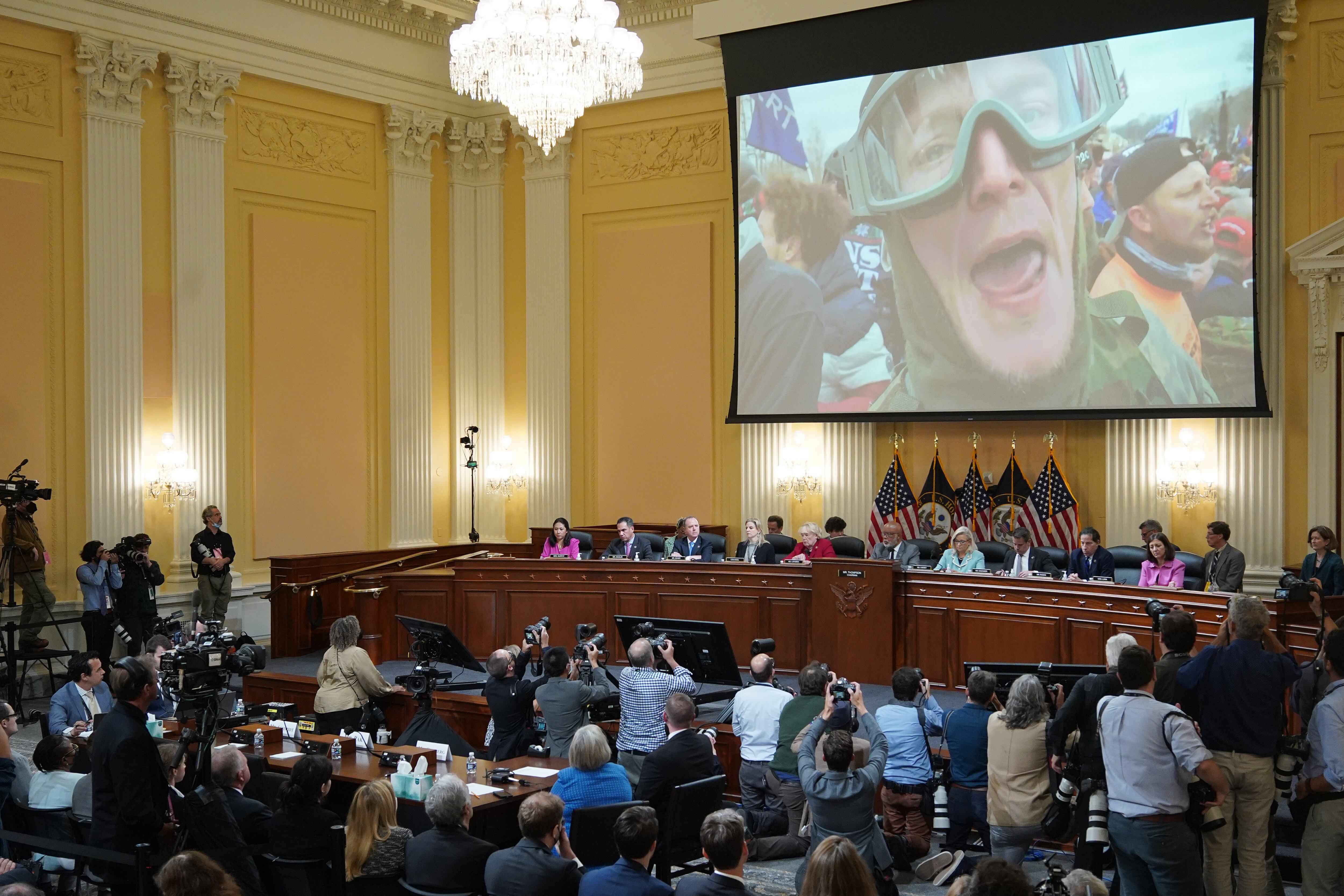 Members of Congress and the media look on as the face of an insurrectionist plays on a large screen