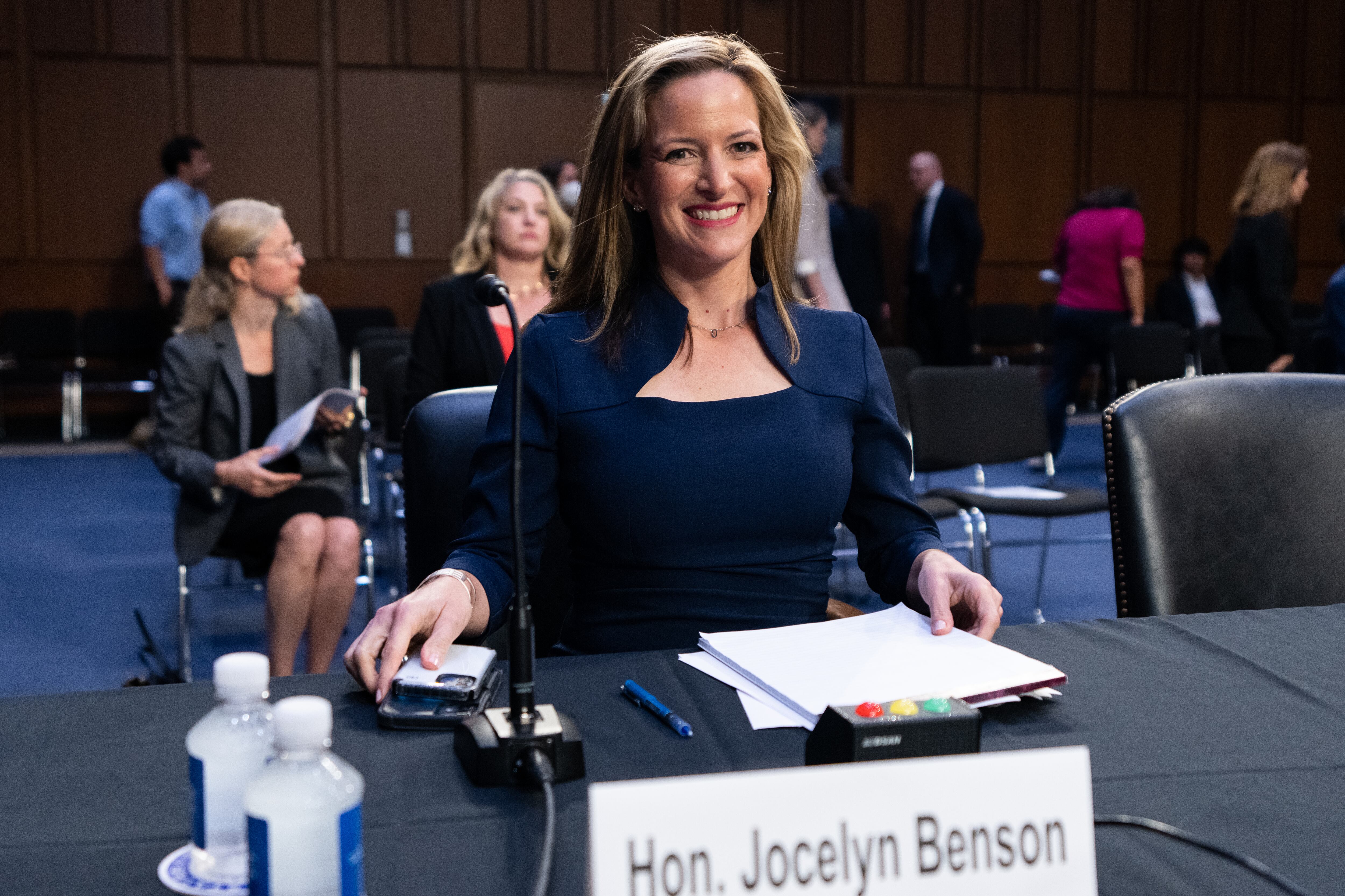 A woman seated at a table in a hearing room smiles. A placard reads “Hon. Jocelyn Benson.”