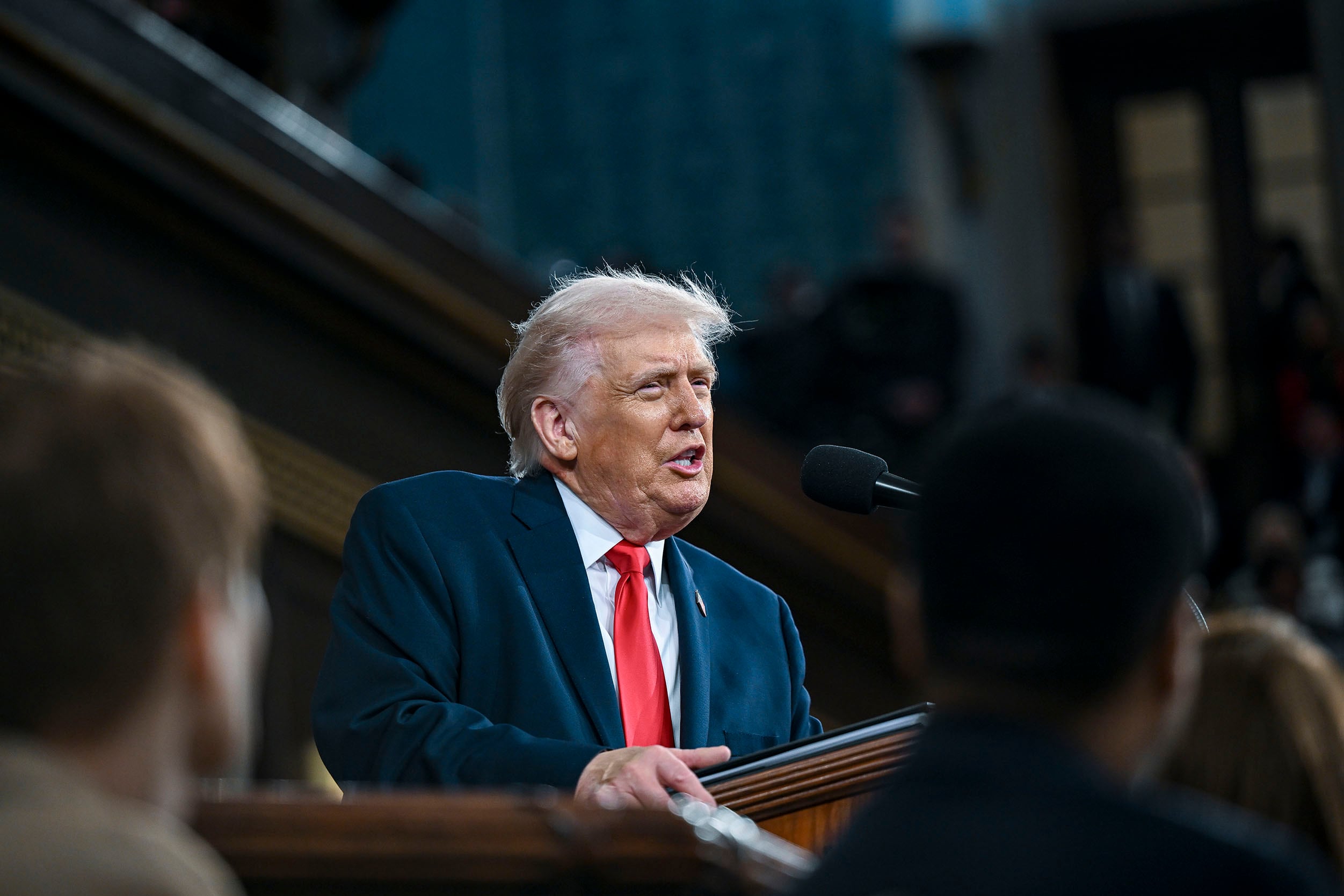 A photograph of President Donal Trump in a blue suit and red tie speaking into a microphone with a few people in the foreground.
