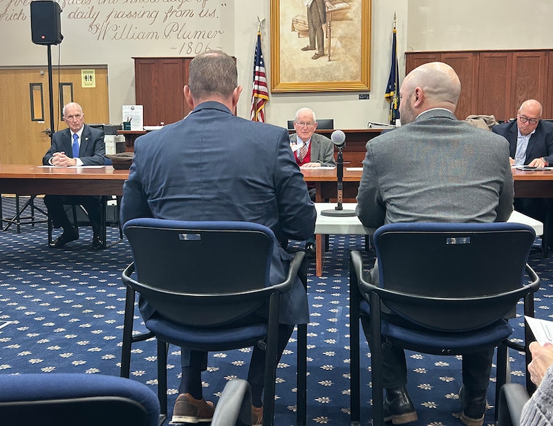 Men in suits sit at tables in a room with blue carpet.