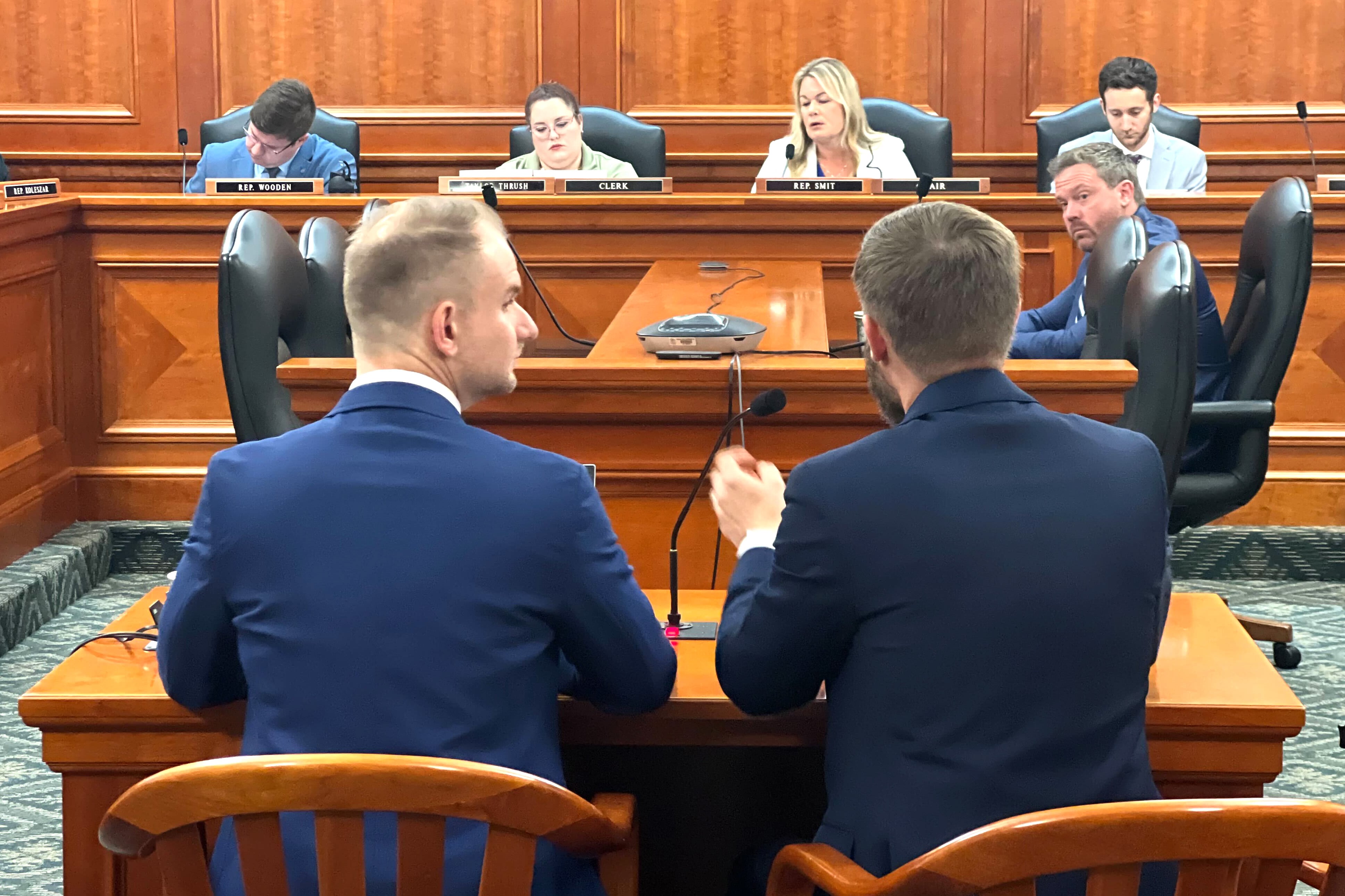 Two men in navy suits sit before people in front of the room.