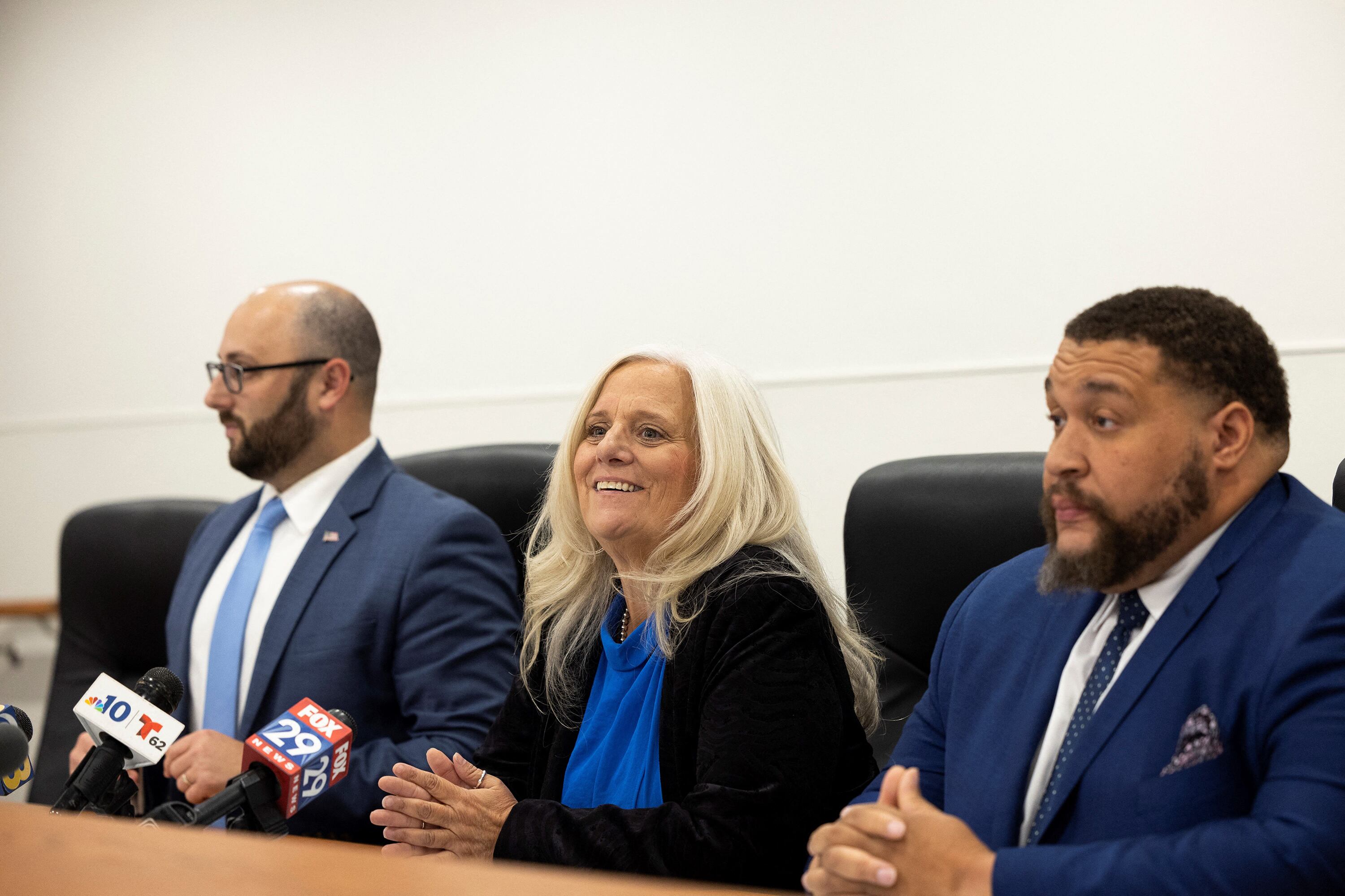 Three people wearing business clothes sit in a row at a wooden desk with a white wall in the background.