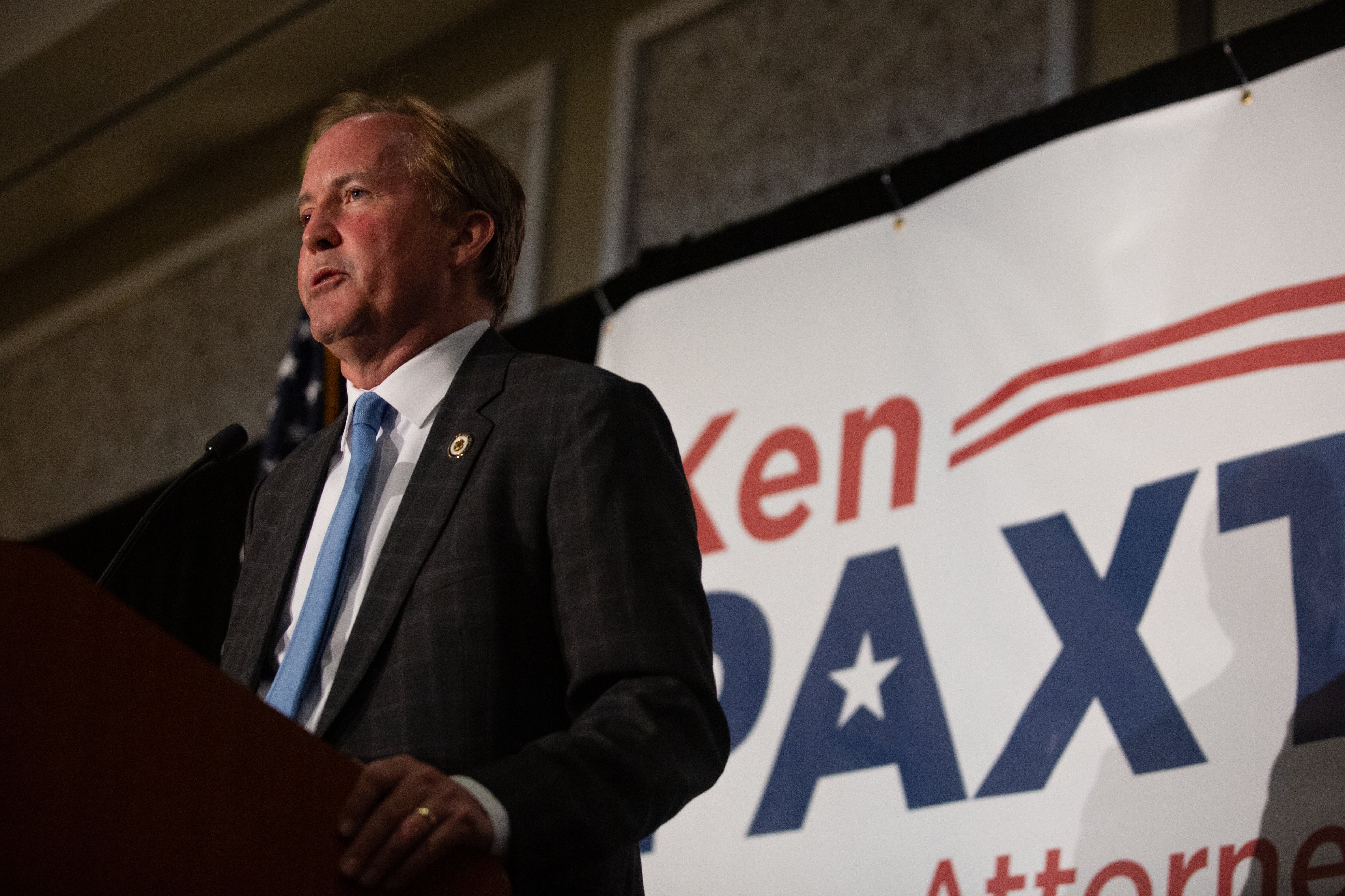 man stands at a lectern before a campaign sign reading “Ken Paxton”