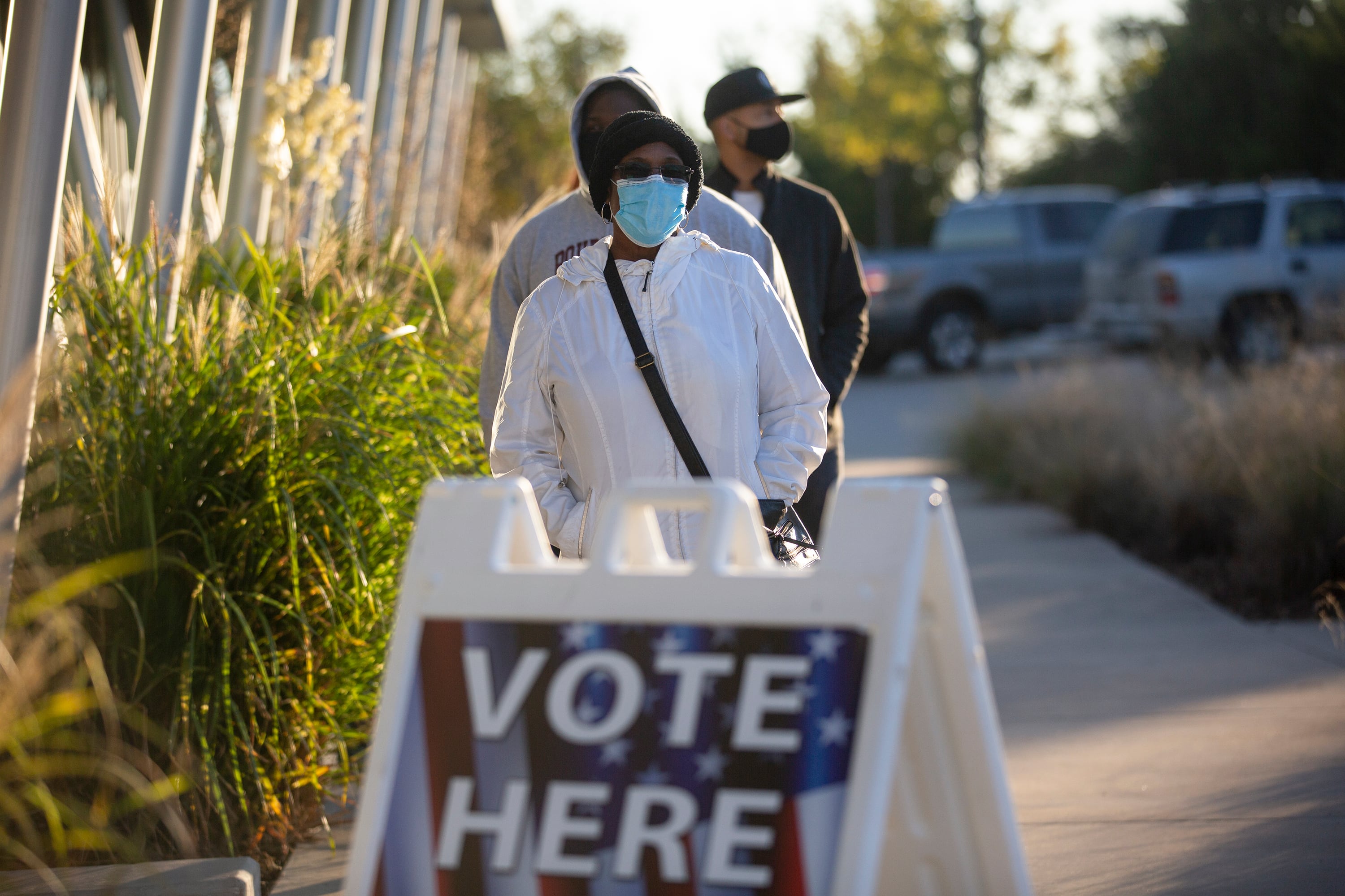 A line of people is formed on a sidewalk along a building. In front of the line there's a sign that says "vote here"