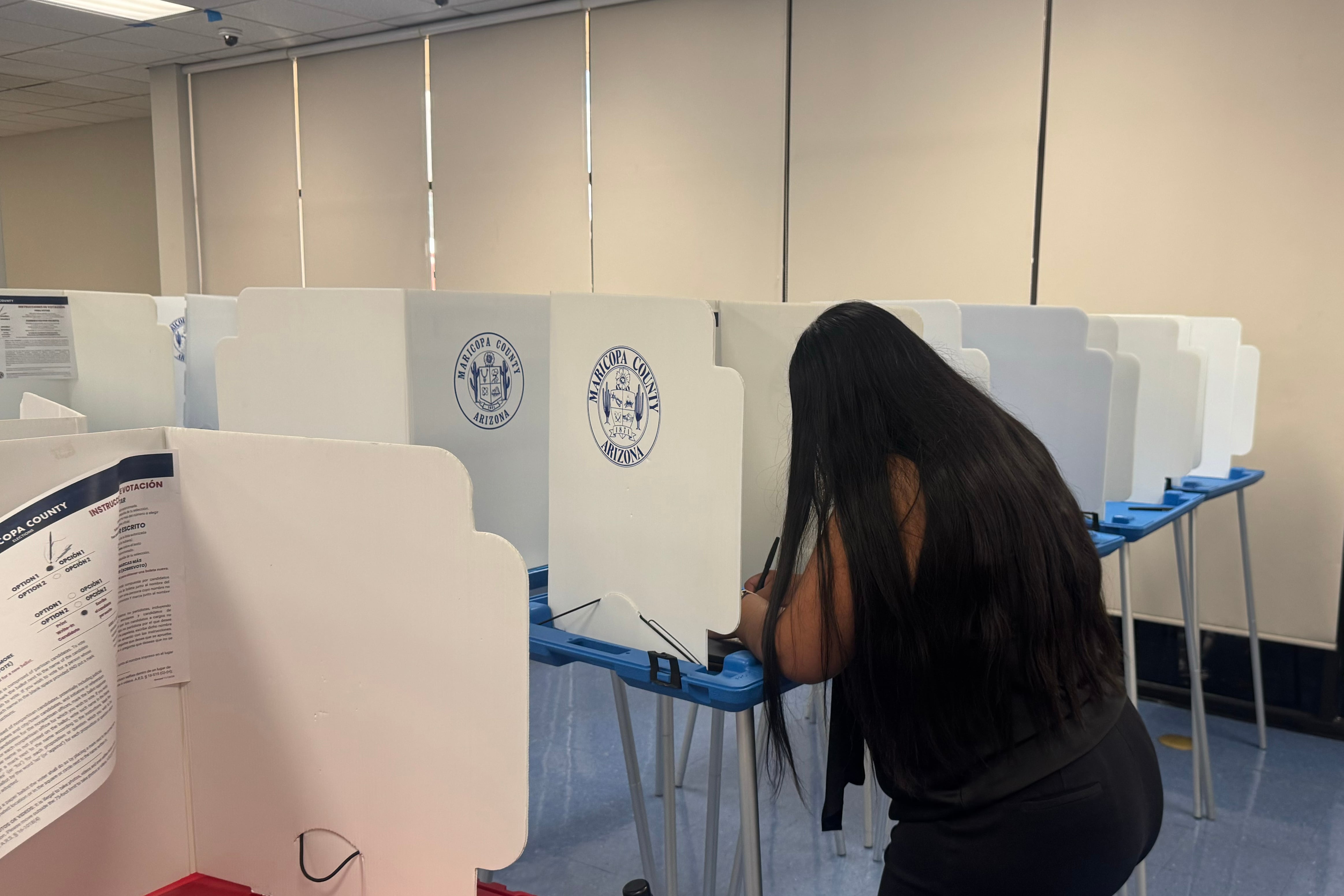 A photograph of a woman with long dark hair at a voting booth in a room.