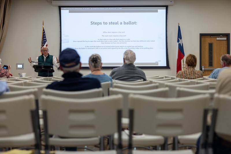 A person stands at a podium next to a projector that reads "Steps to steal a ballot" with a couple of people in chairs in rows.