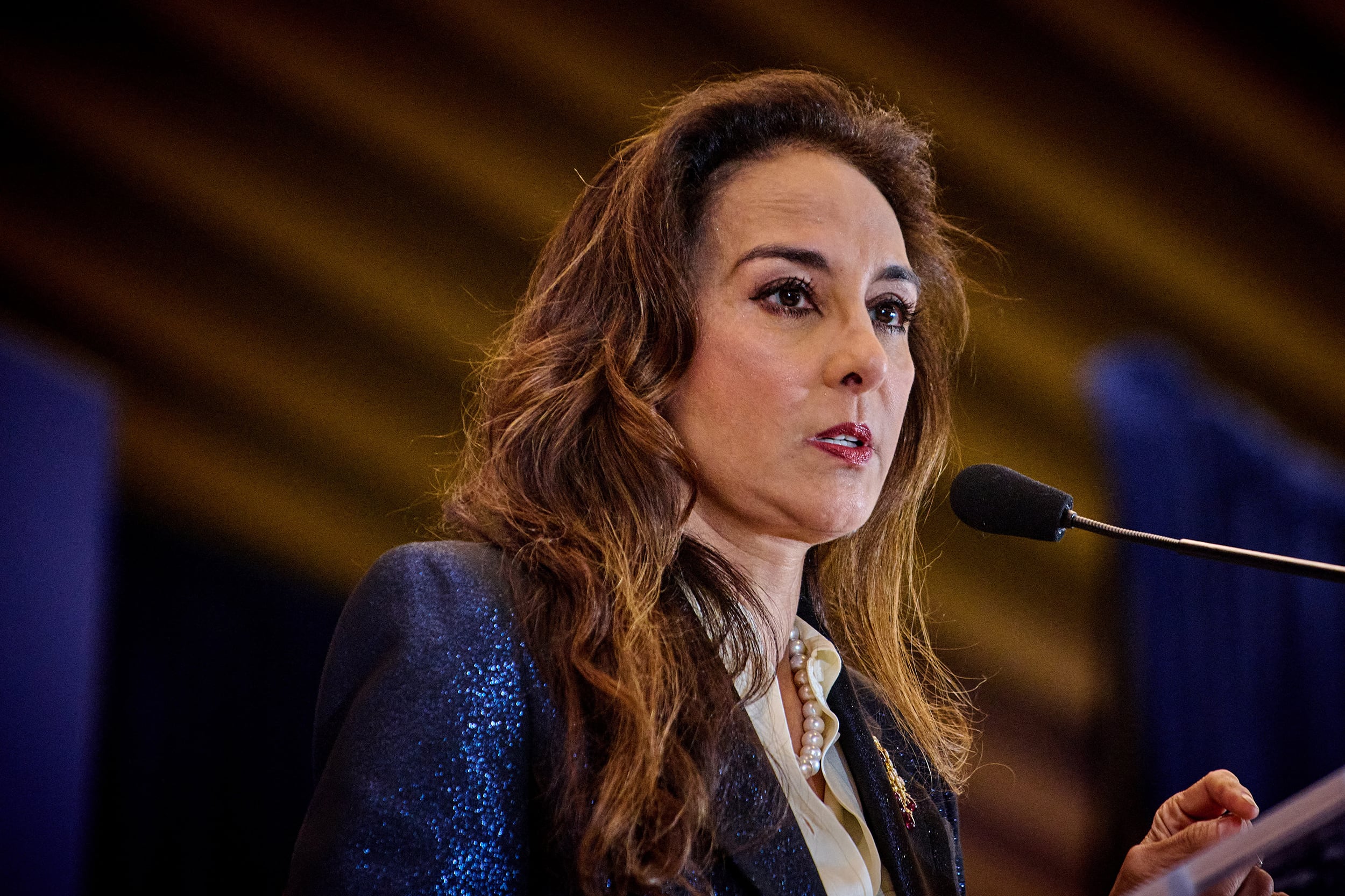 A photograph of a woman with long dark hair in a suit speaking from behind a microphone.