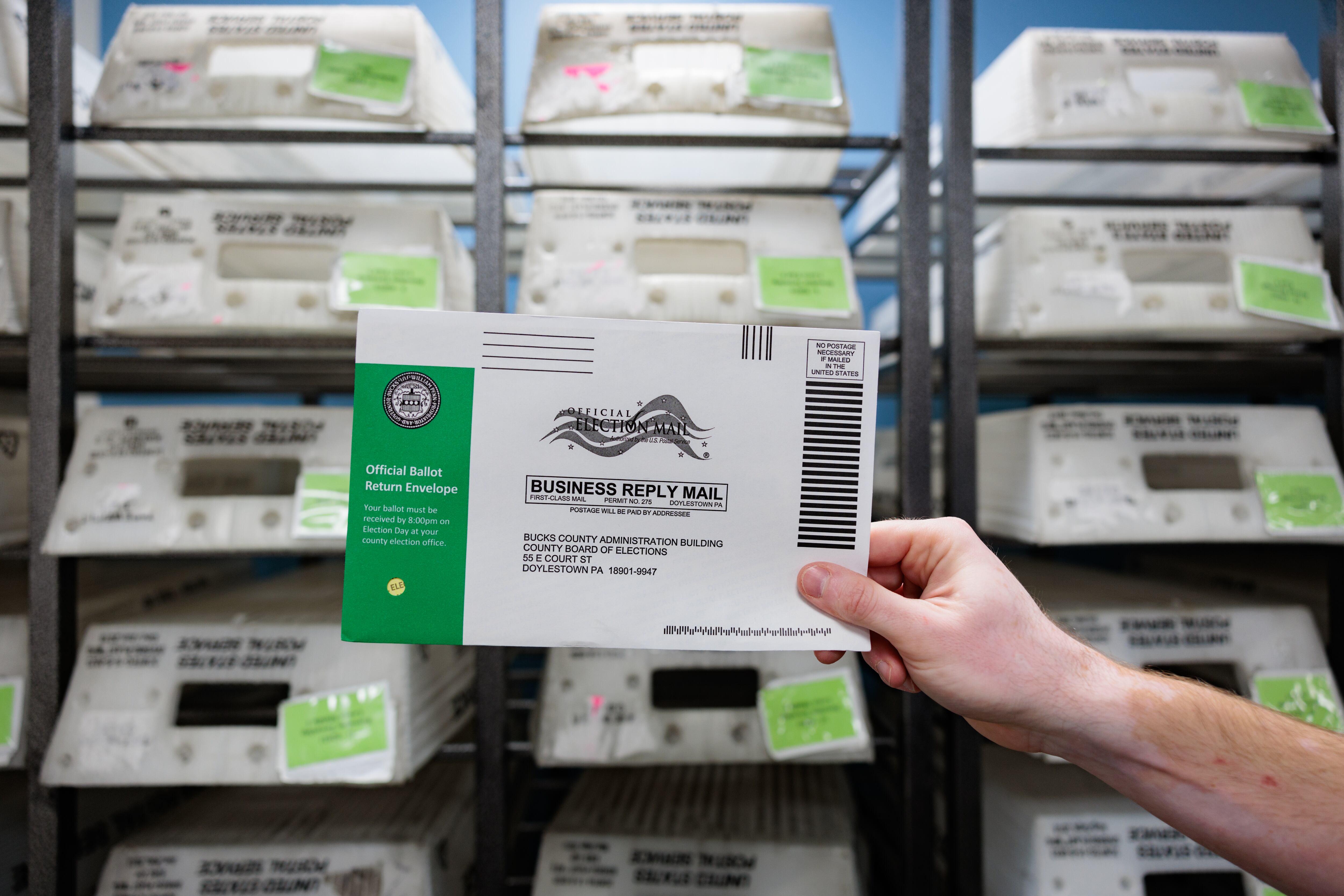 A hand holds up an envelope with a green stripe against a background of shelves of plastic mail bins