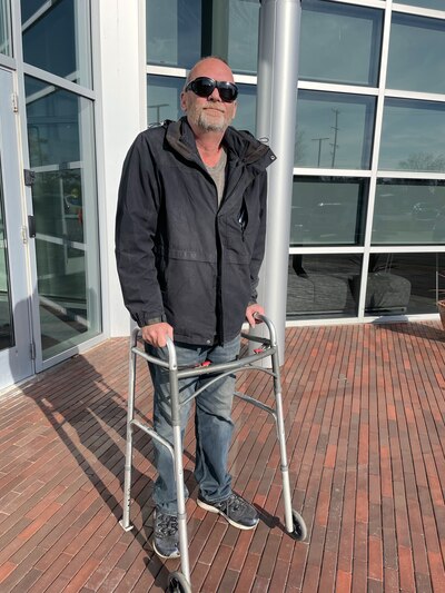 A man wearing sunglasses, a black jacket and using a walker, poses for a portrait on a red brick walkway with windows in the background.