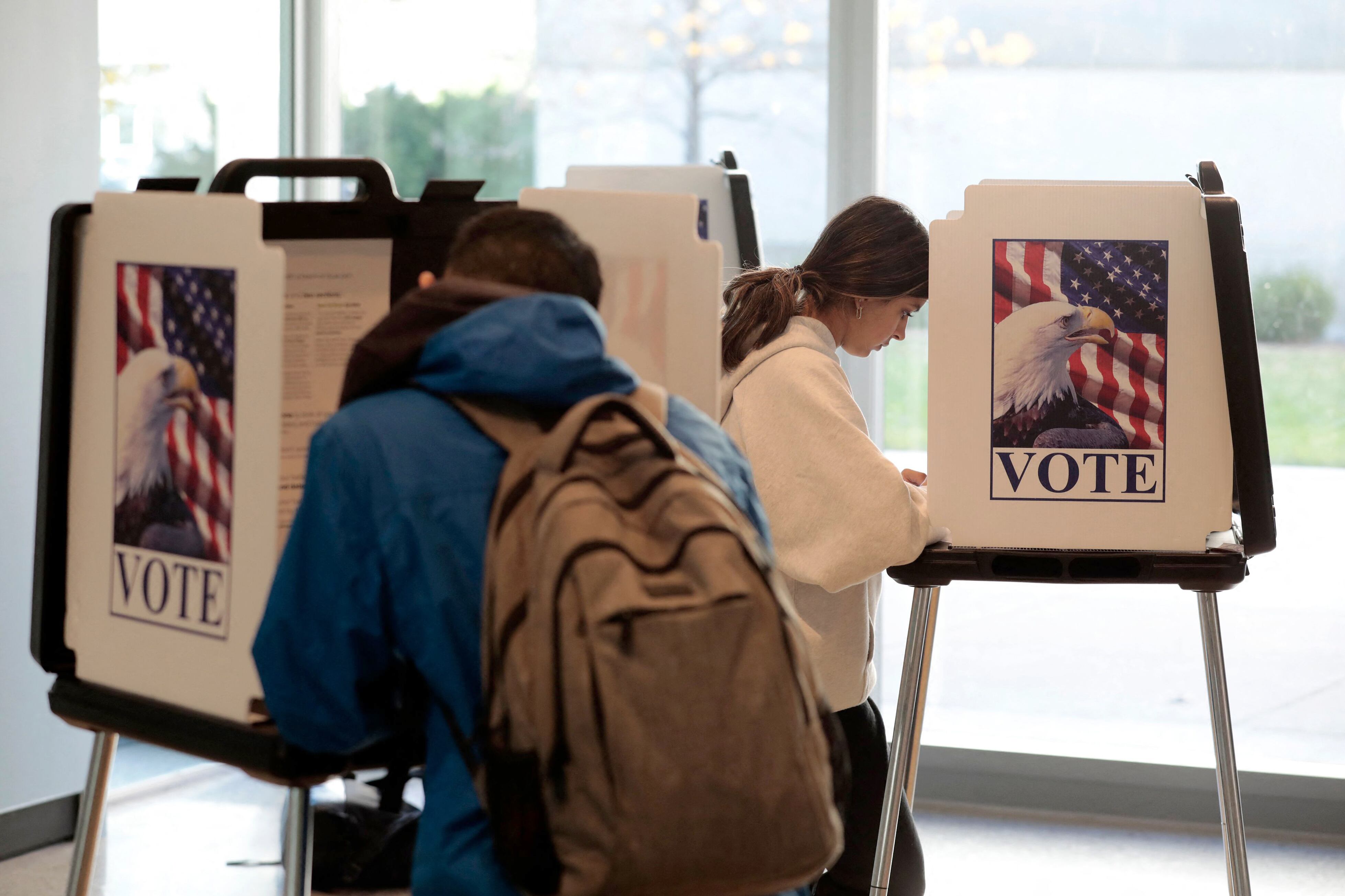 A young dark-haired man with a blue hooded jacket and olive backpack leans over a carrel with his back to the camera. Beyond him, a young dark-haired woman in a white hooded sweatshirt leans over a different carrel, clearly reading something. 