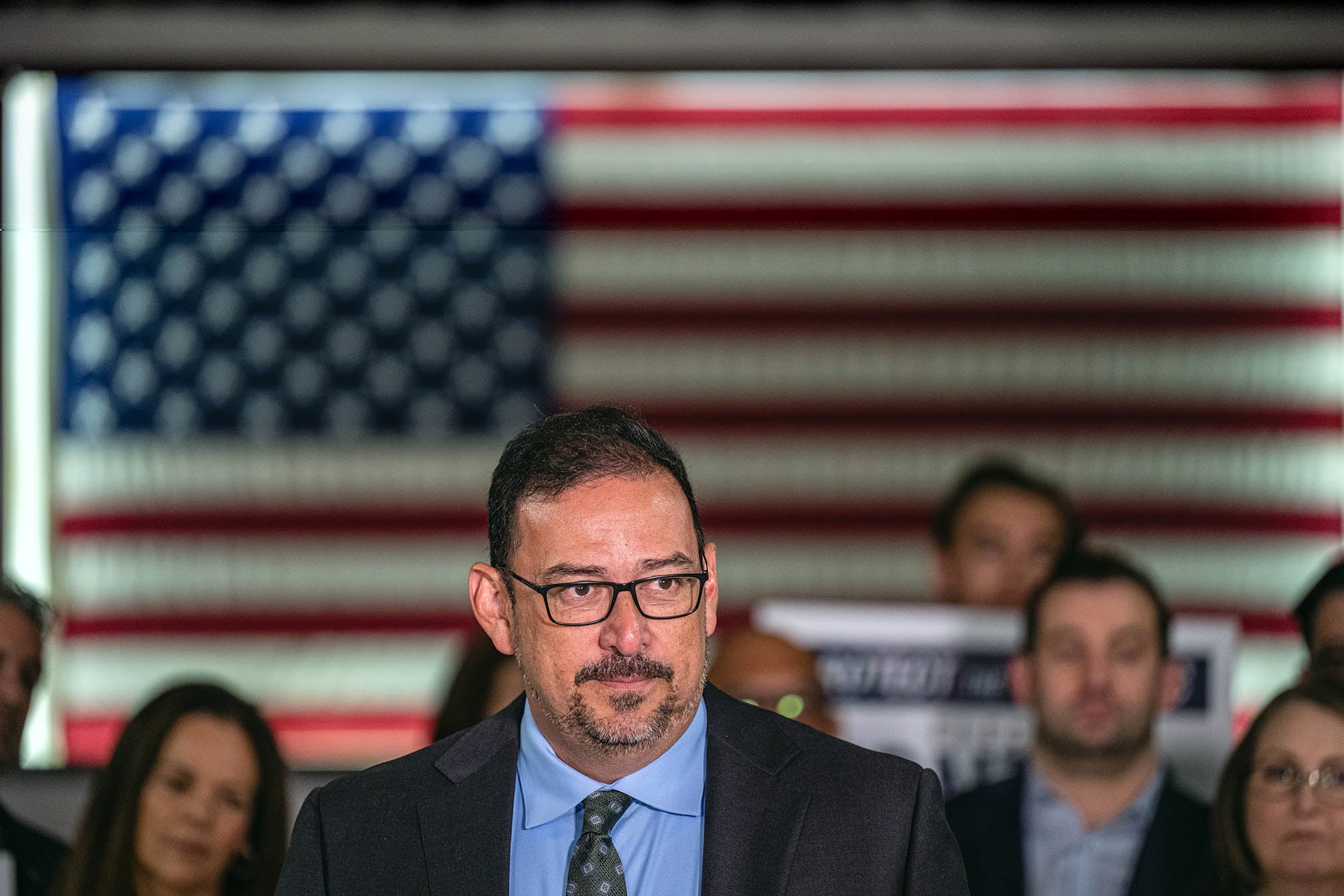 A man with short dark hair and wearing a dark suit stands in front of an American flag and a group of people.