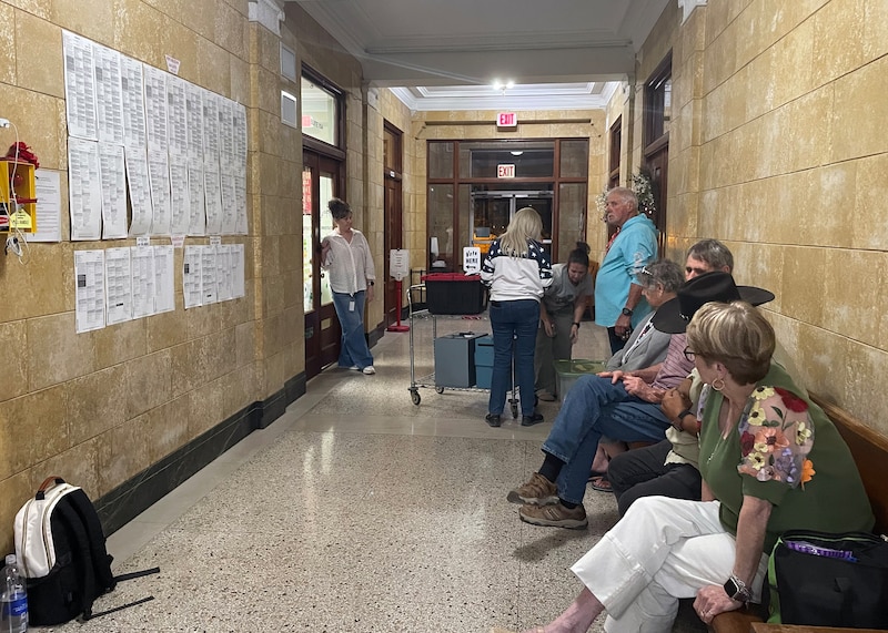 A photograph of a hallway with one side lined with people. some are sitting and some are standing.