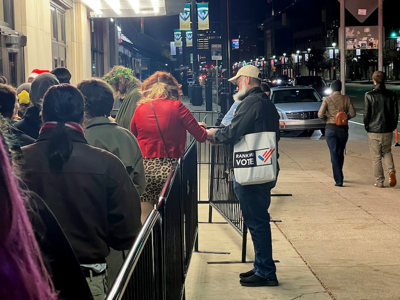 A photograph of a main holding a tote bag speaking to a person in a red sweater standing in a line outside.