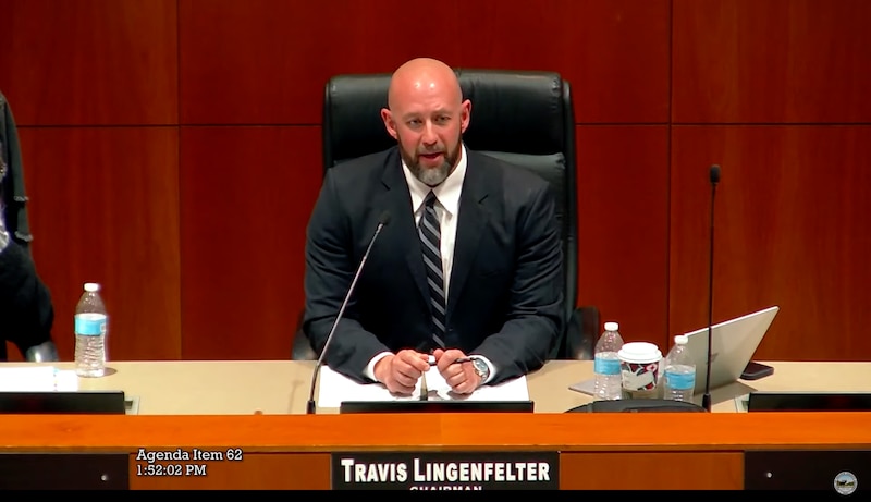 a man wearing a suit sits behind a podium with his name across.