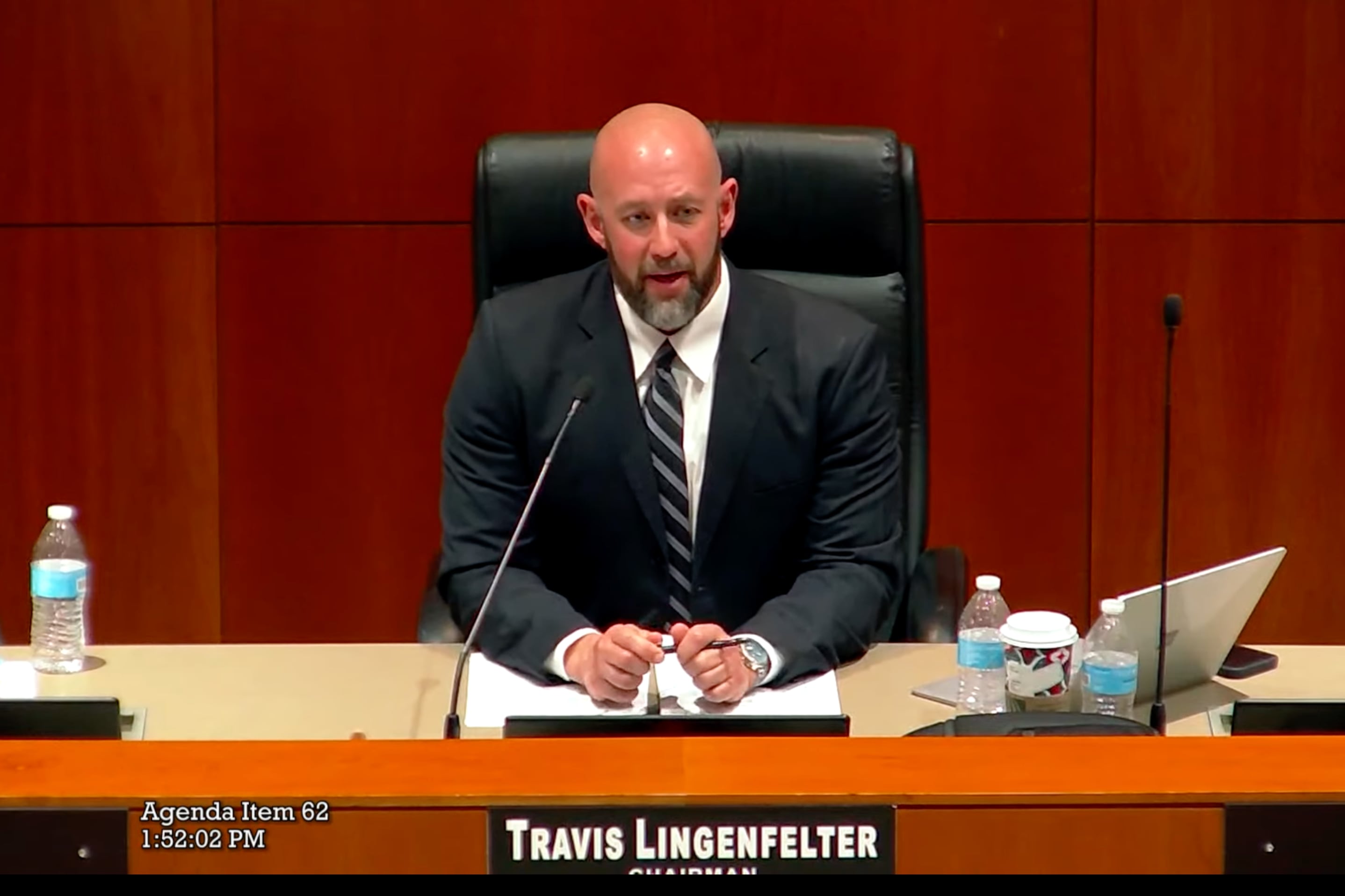 a man wearing a suit sits behind a podium with his name across.