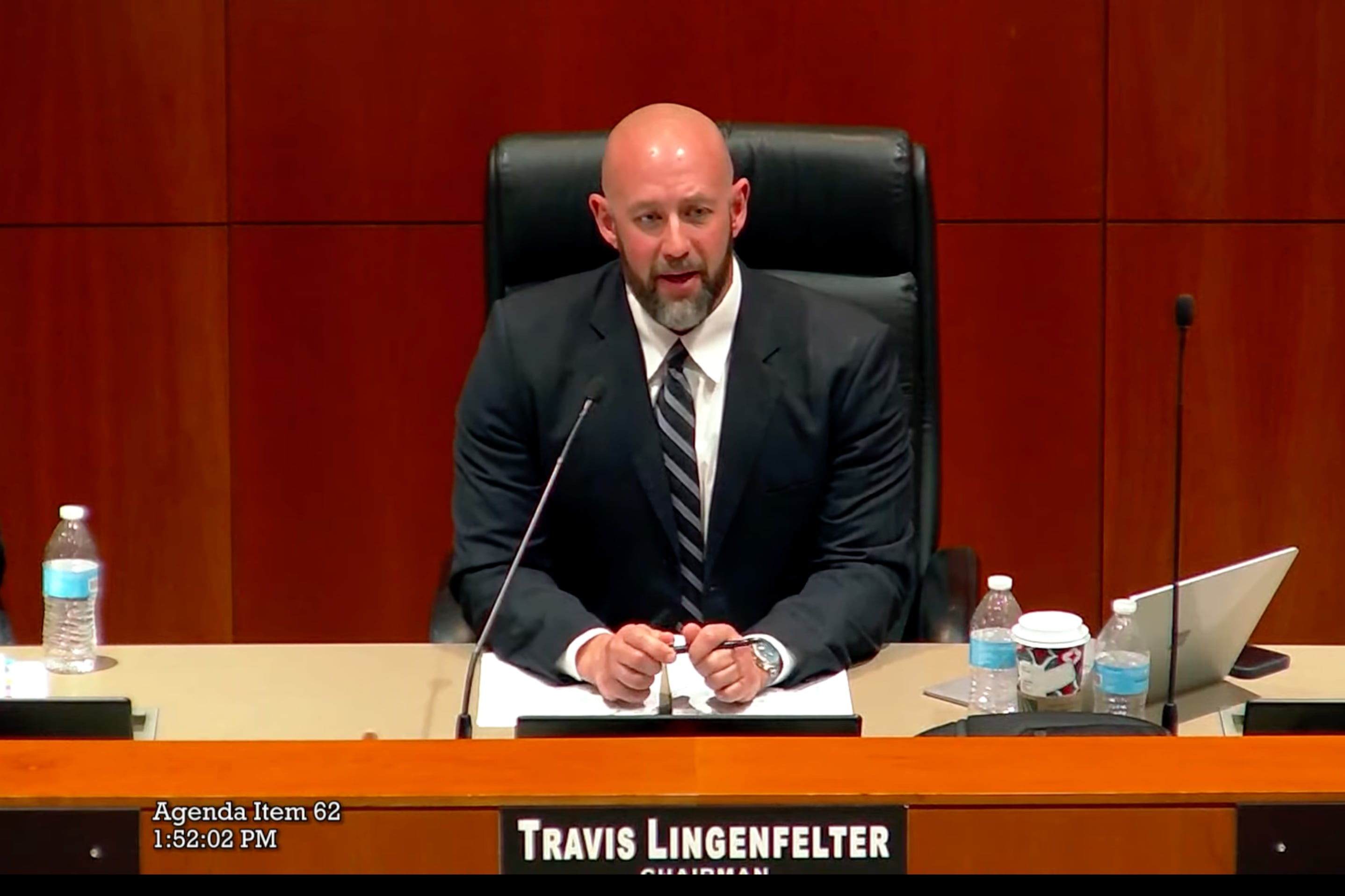 a man wearing a suit sits behind a podium with his name across.