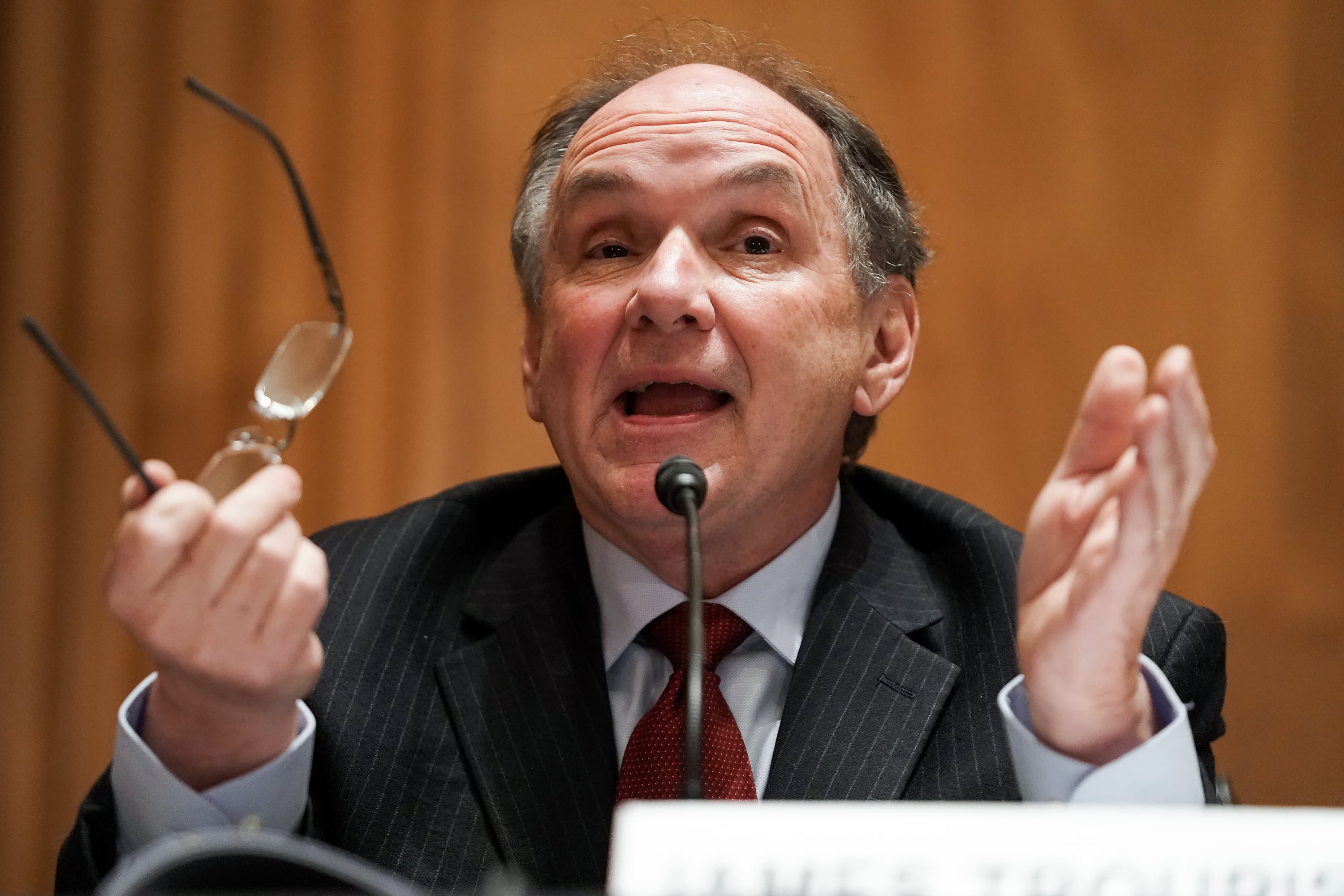 A man in a dark suit holds his classes while talking at a table.