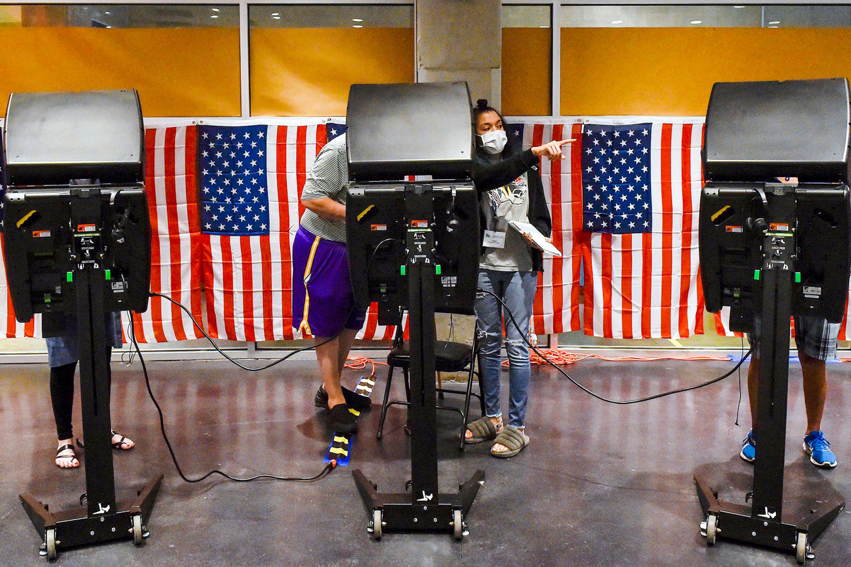 Four people stand behind three large black voting machines evenly spread out in front of a wall of American flags hanging perfectly in a row.