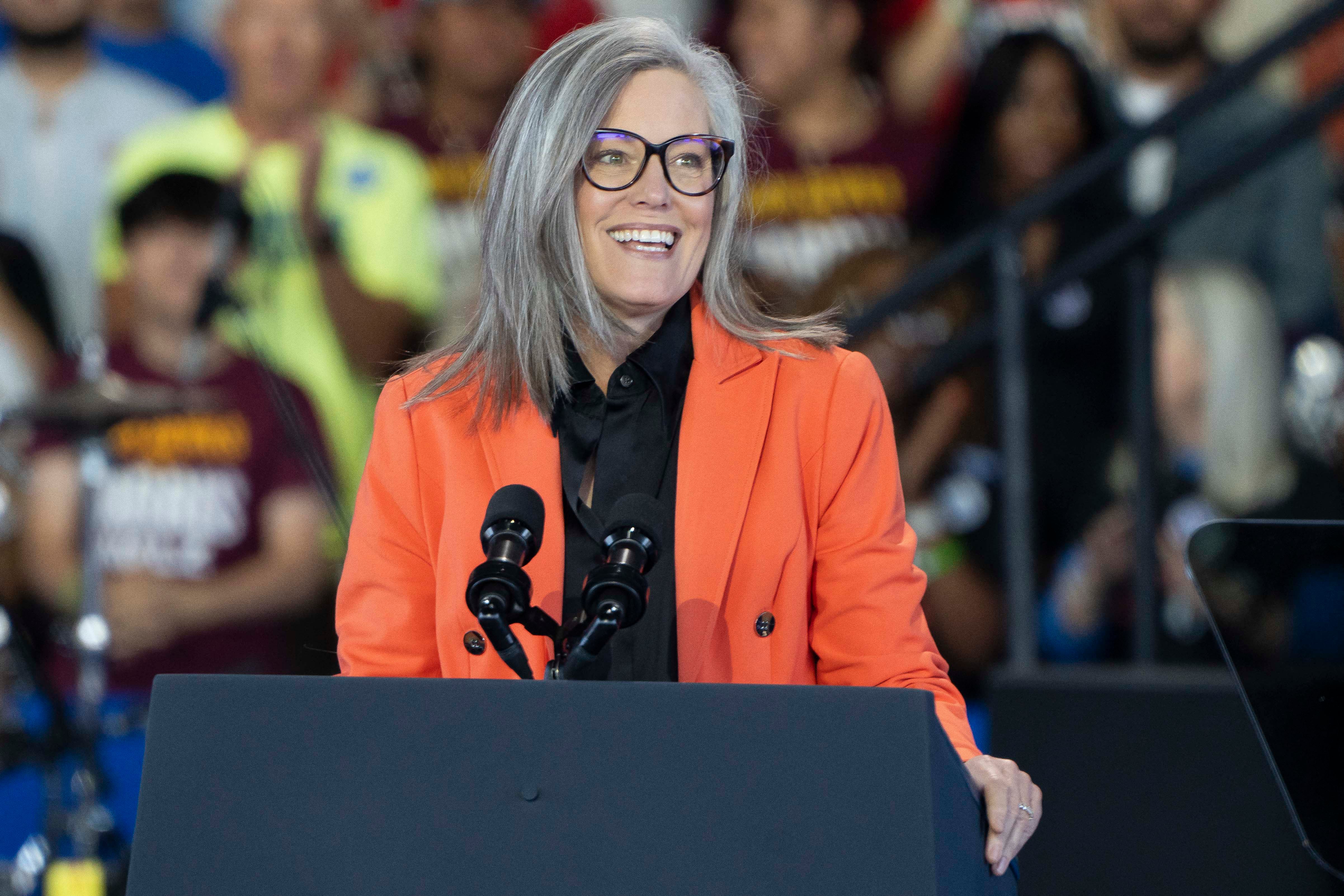 A woman in an orange suit jacket speaks from behind a podium and in front of a crowd sitting in the background.