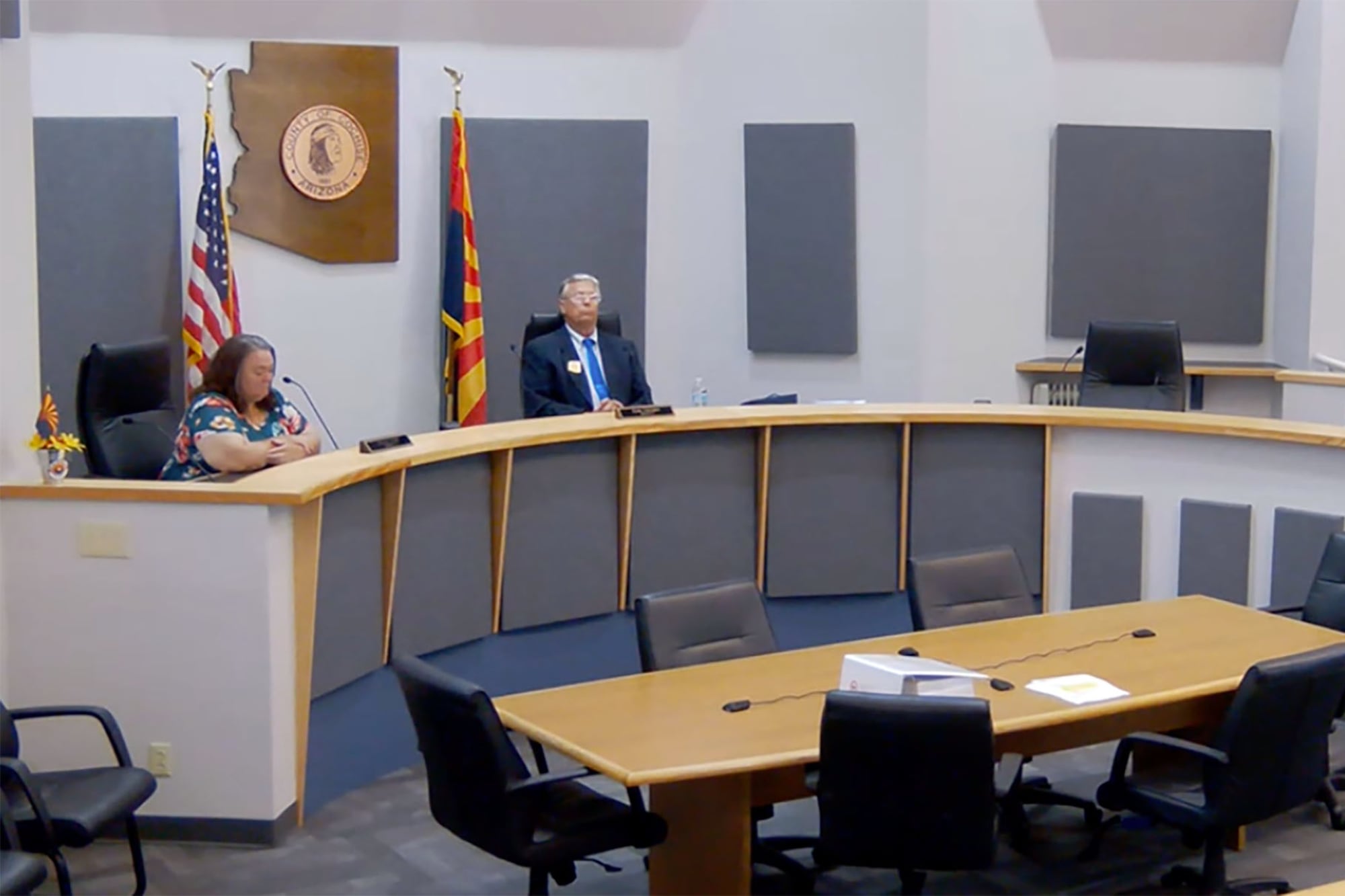 Two adults sit at a rounded wooden table along a wall with an American and Arizona flag in the background.