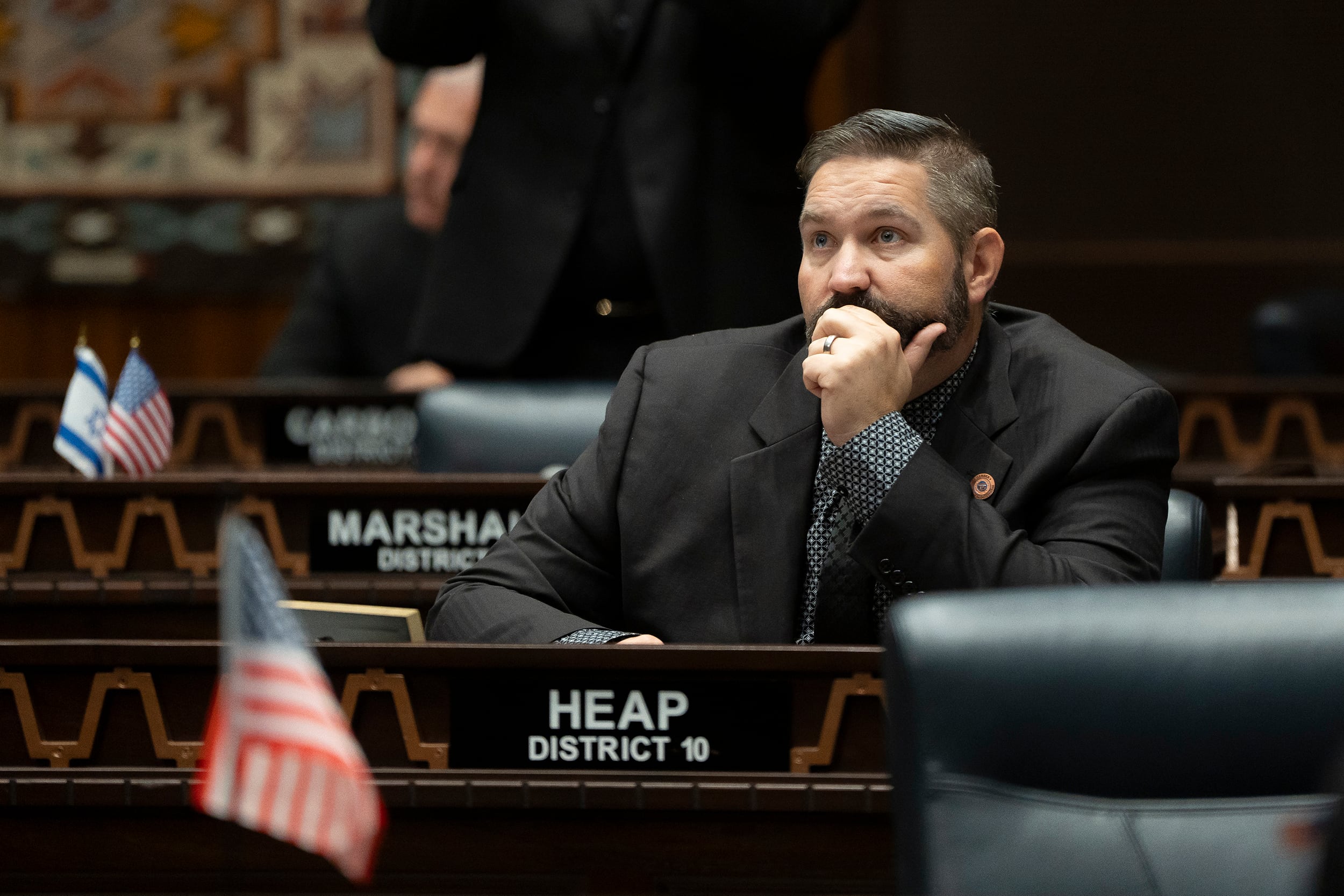 A photograph of a white man in a suit sitting at a wooden desk with his name in the front.