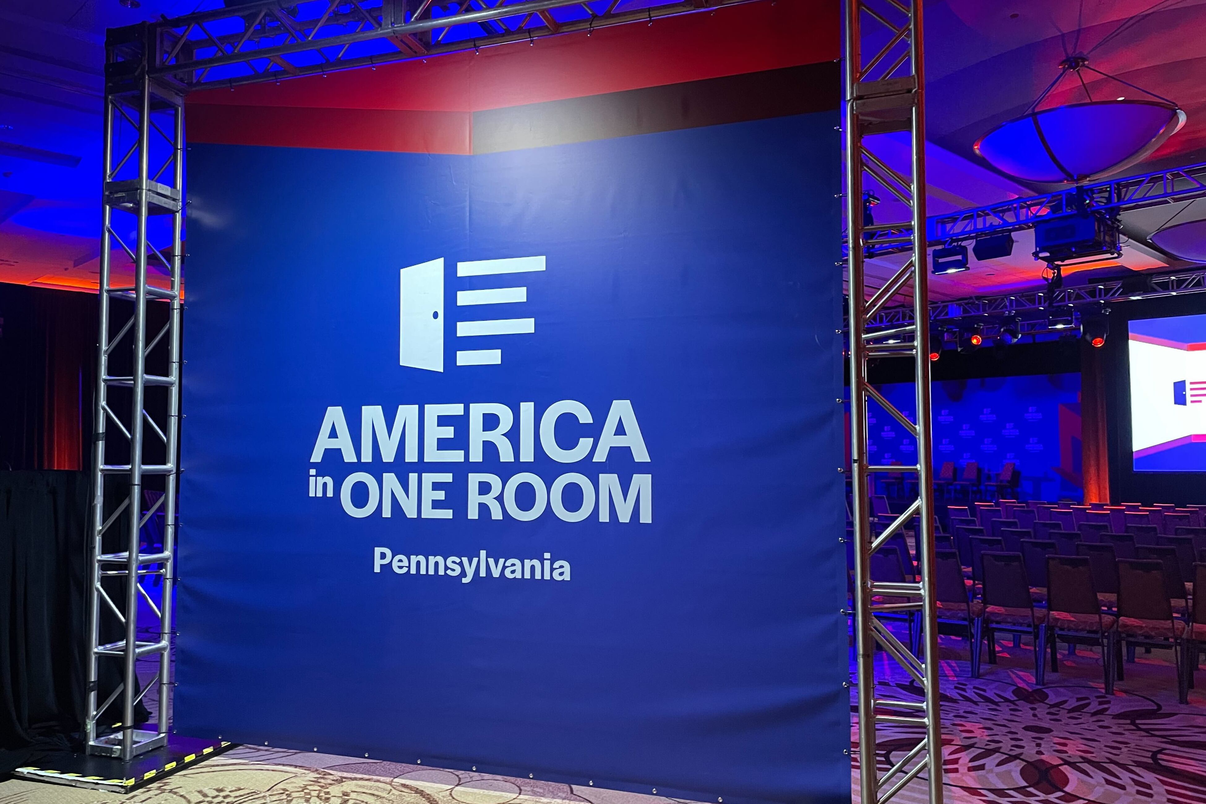 A blue background with white words that read "America in One Room Pennsylvania," with a large theater seating in the background.