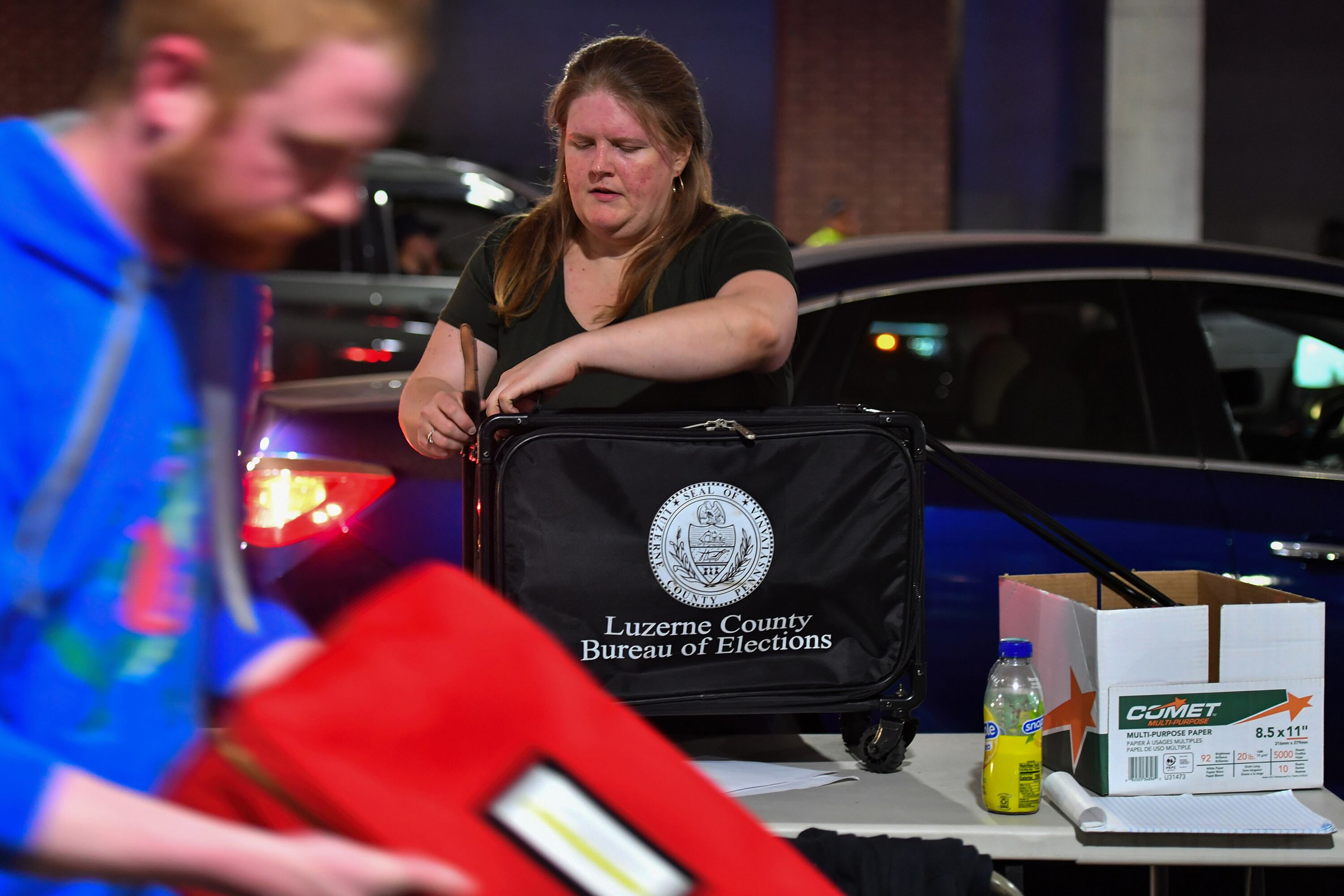 A person with long hair closes up a black box with words and a logo on the side and in the foreground, a person walks by carrying a red box.