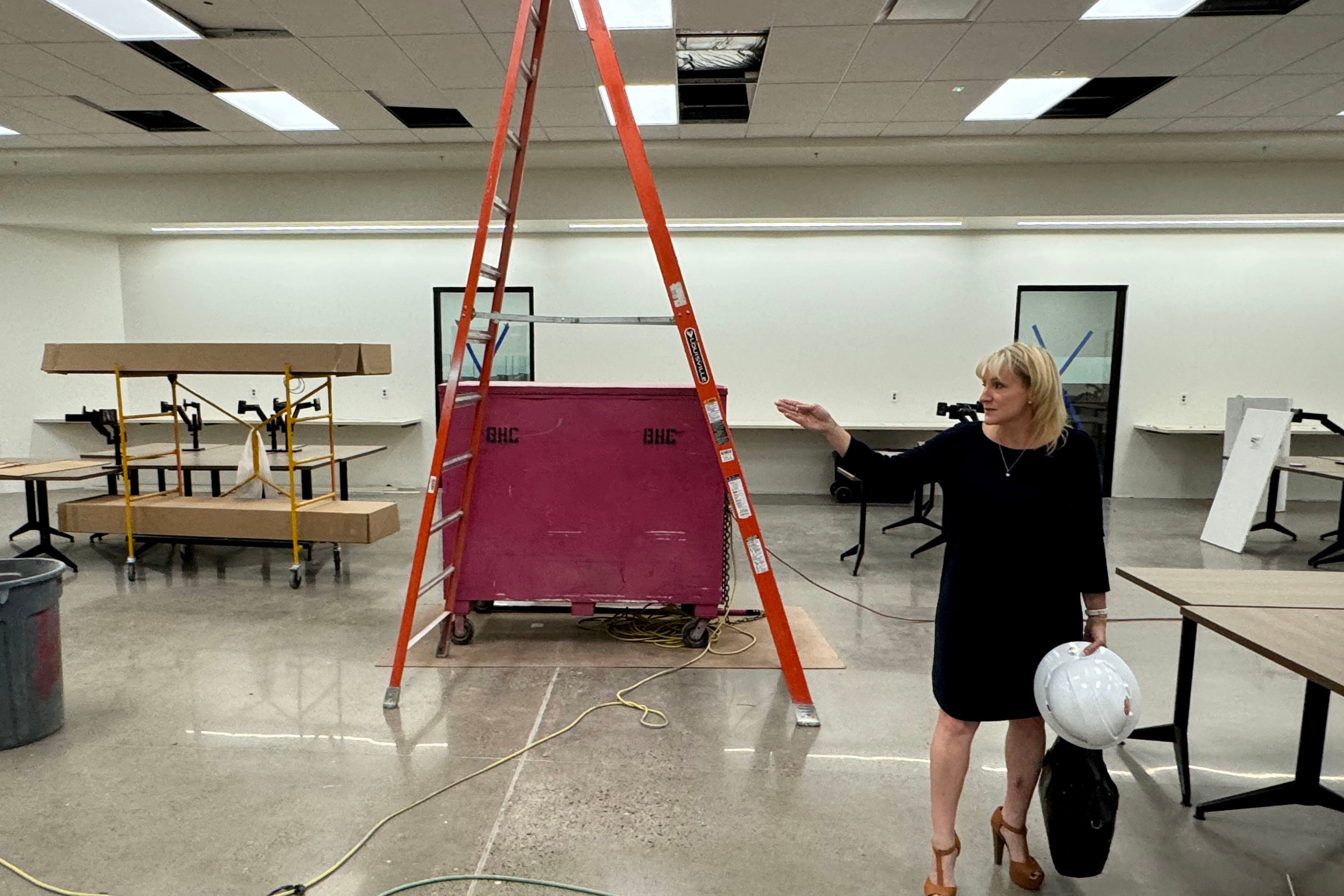 A woman with short blonde hair and wearing a black dress holds a bag and a hard hat while touring a building that is under construction.