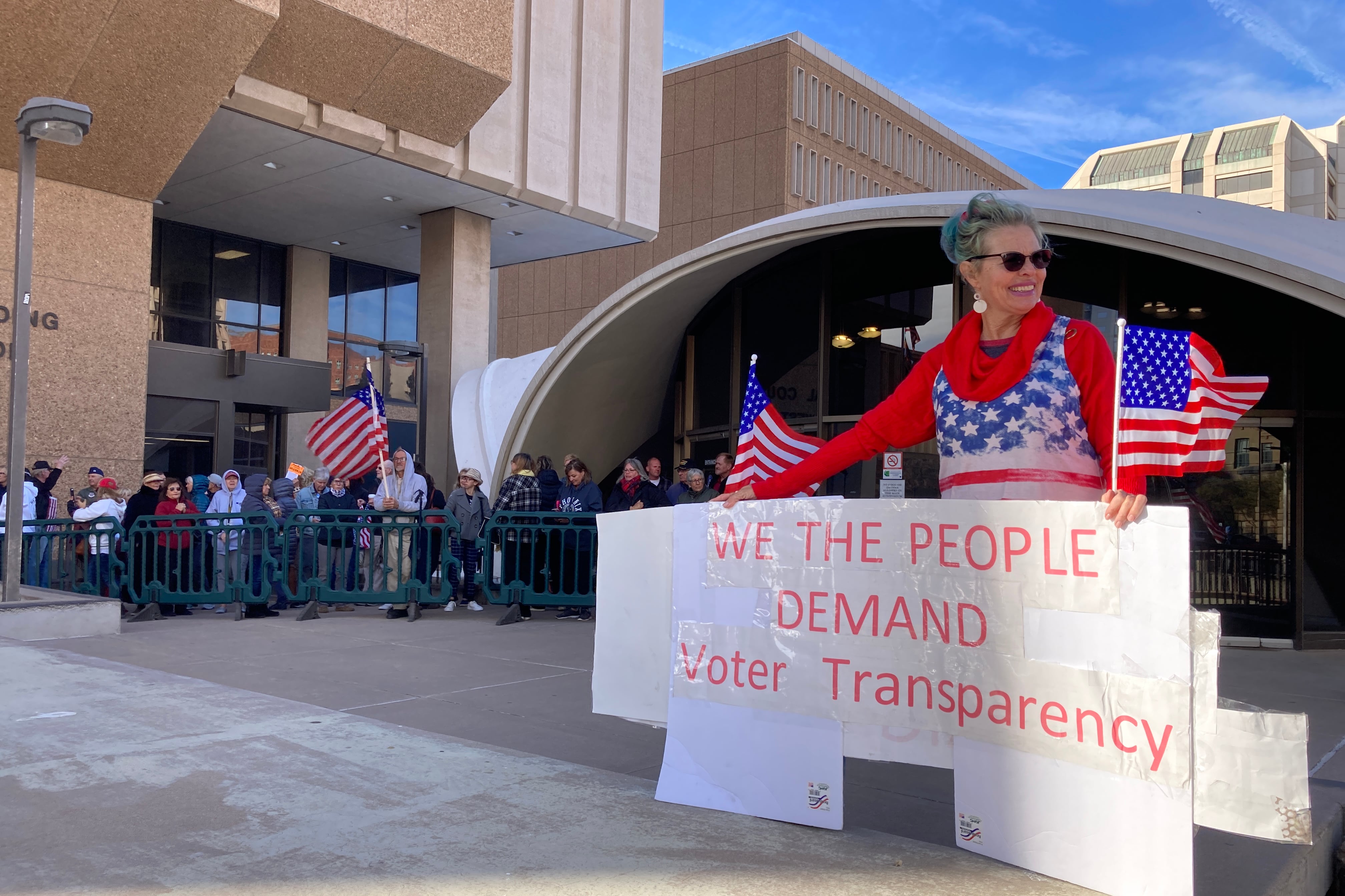 A woman holds a large sign that reads, “We the people demand voter transparency” while people line up behind her. 