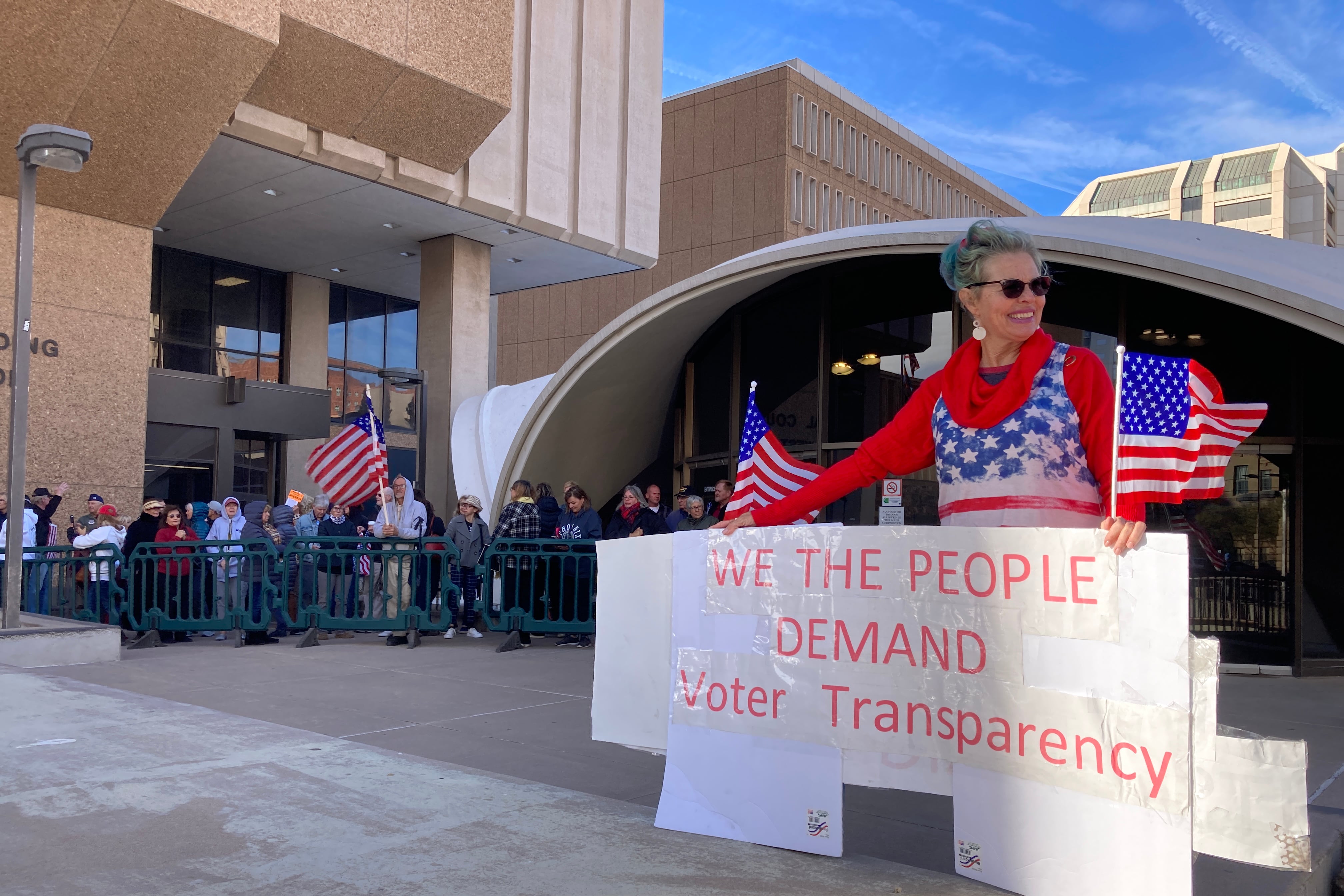 A woman holds a large sign that reads, “We the people demand voter transparency” while people line up behind her.