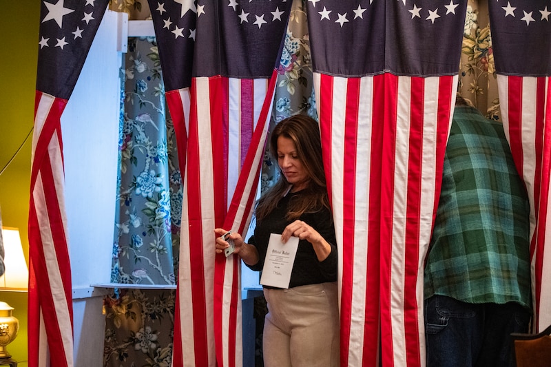 A woman holds a ballot as she walks out of a voting booth with an American flag curtain.