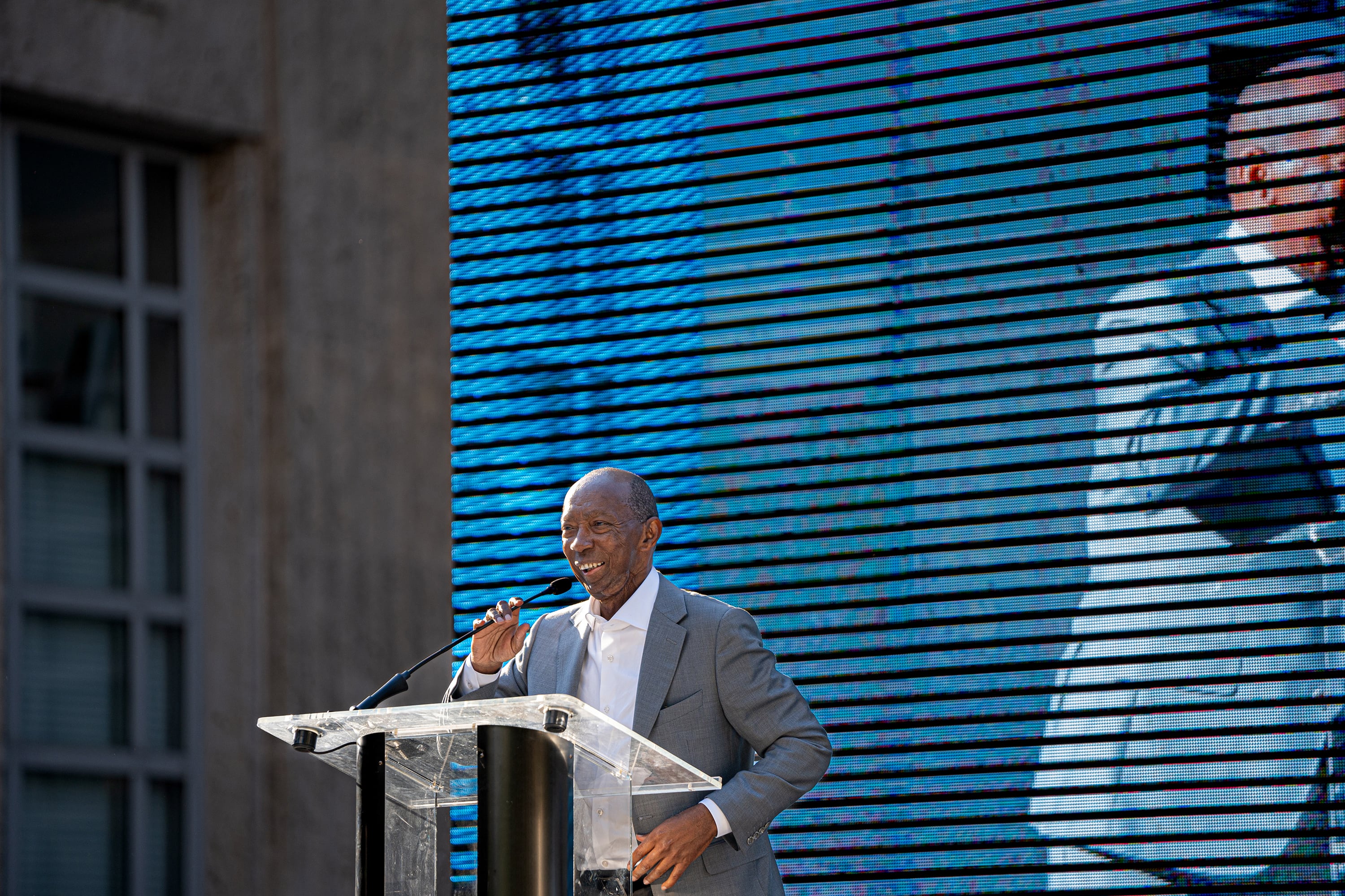 A photograph of a Black man in a suit on a stage with a blue background.