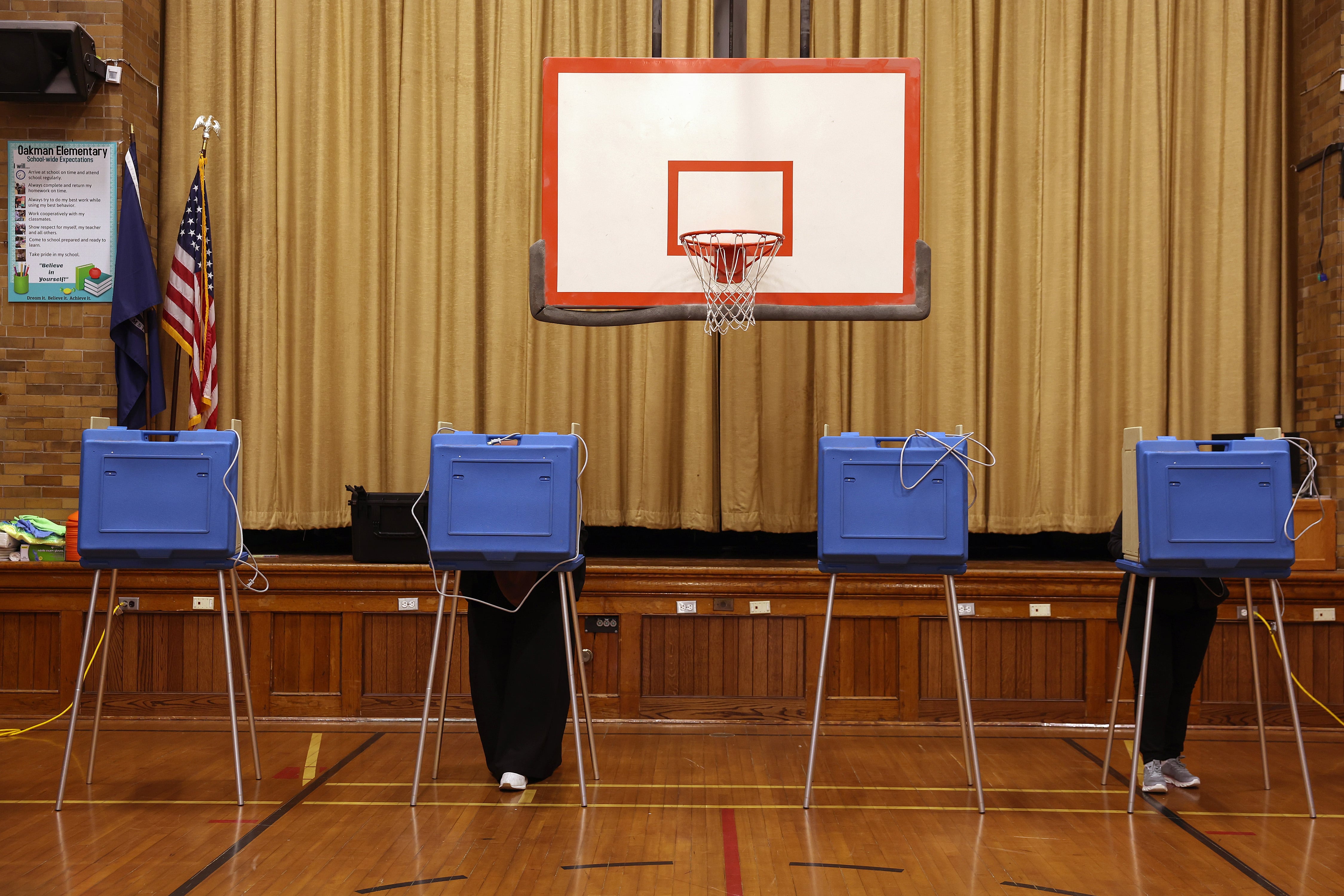 Four blue voter booths lined up in front of a basketball hoop in a school gym.
