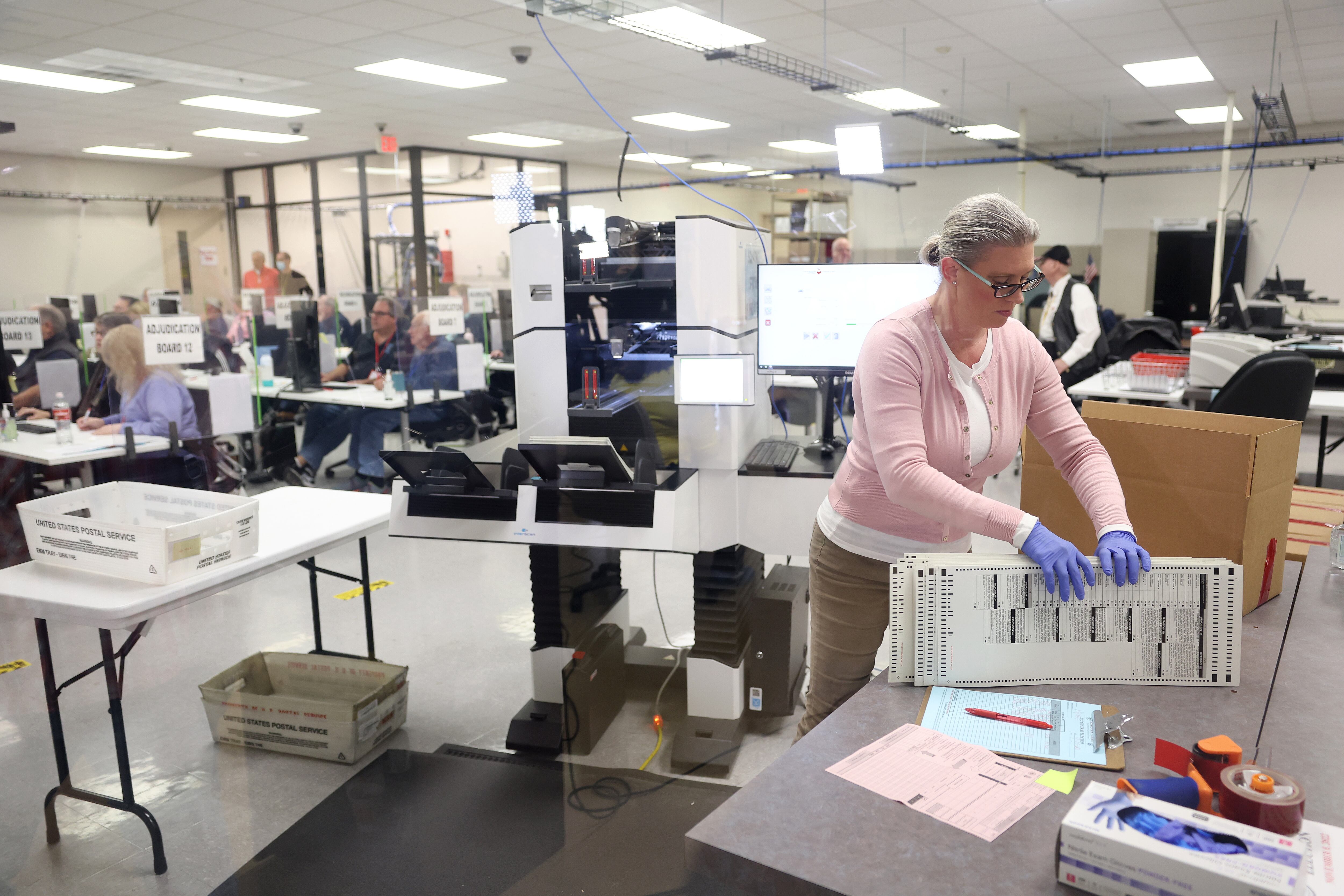 A woman lifts a stack of ballots off a table to load into a large machine behind her
