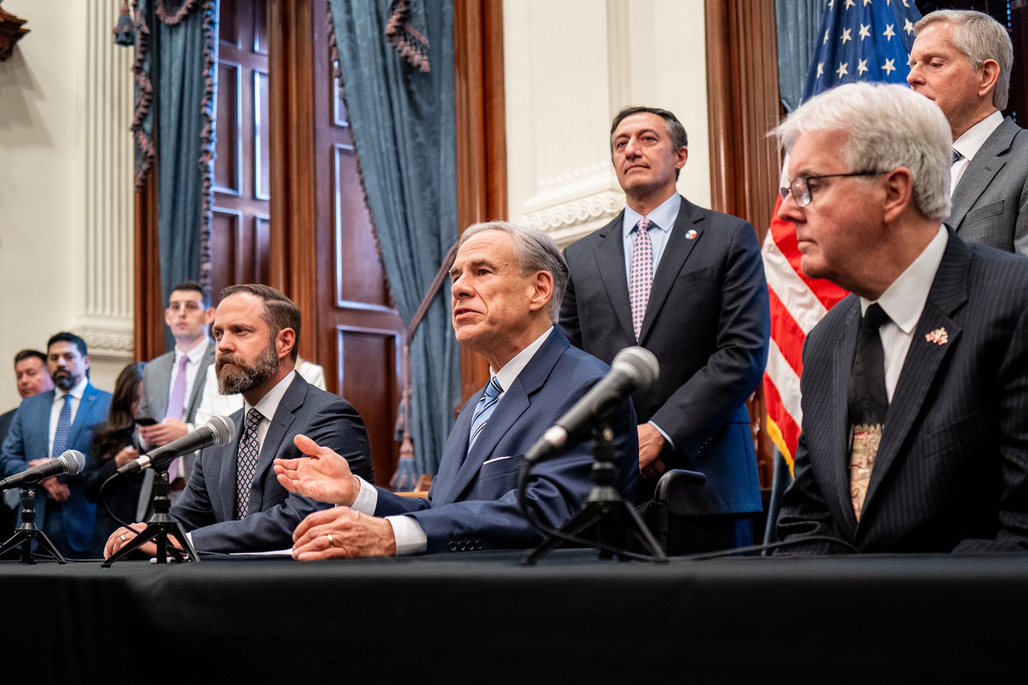 A photograph of a row of white men in suits sitting at a table and a row of men in suits standing in the background.