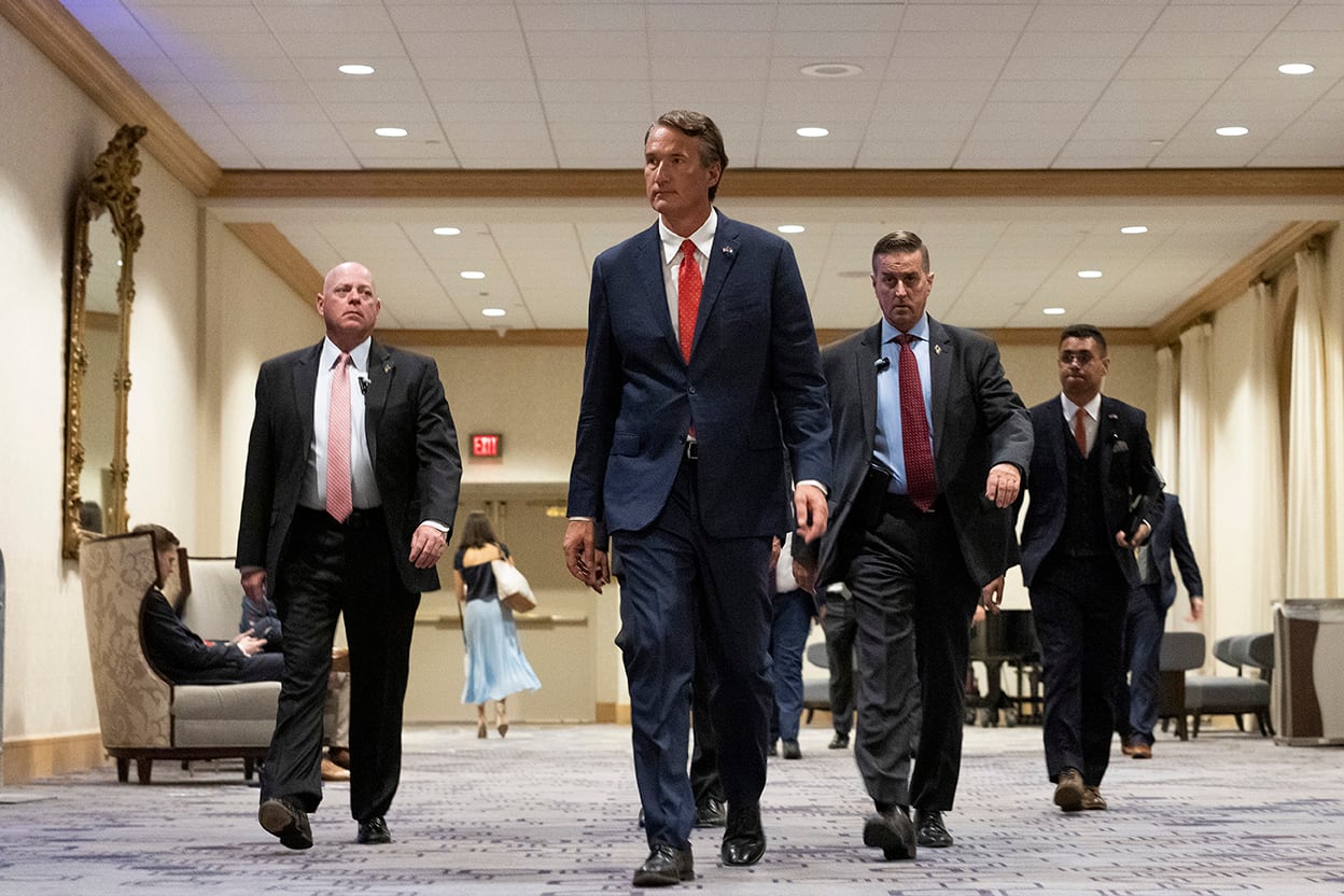 Four men in dark suites and red ties walk down a hallway.