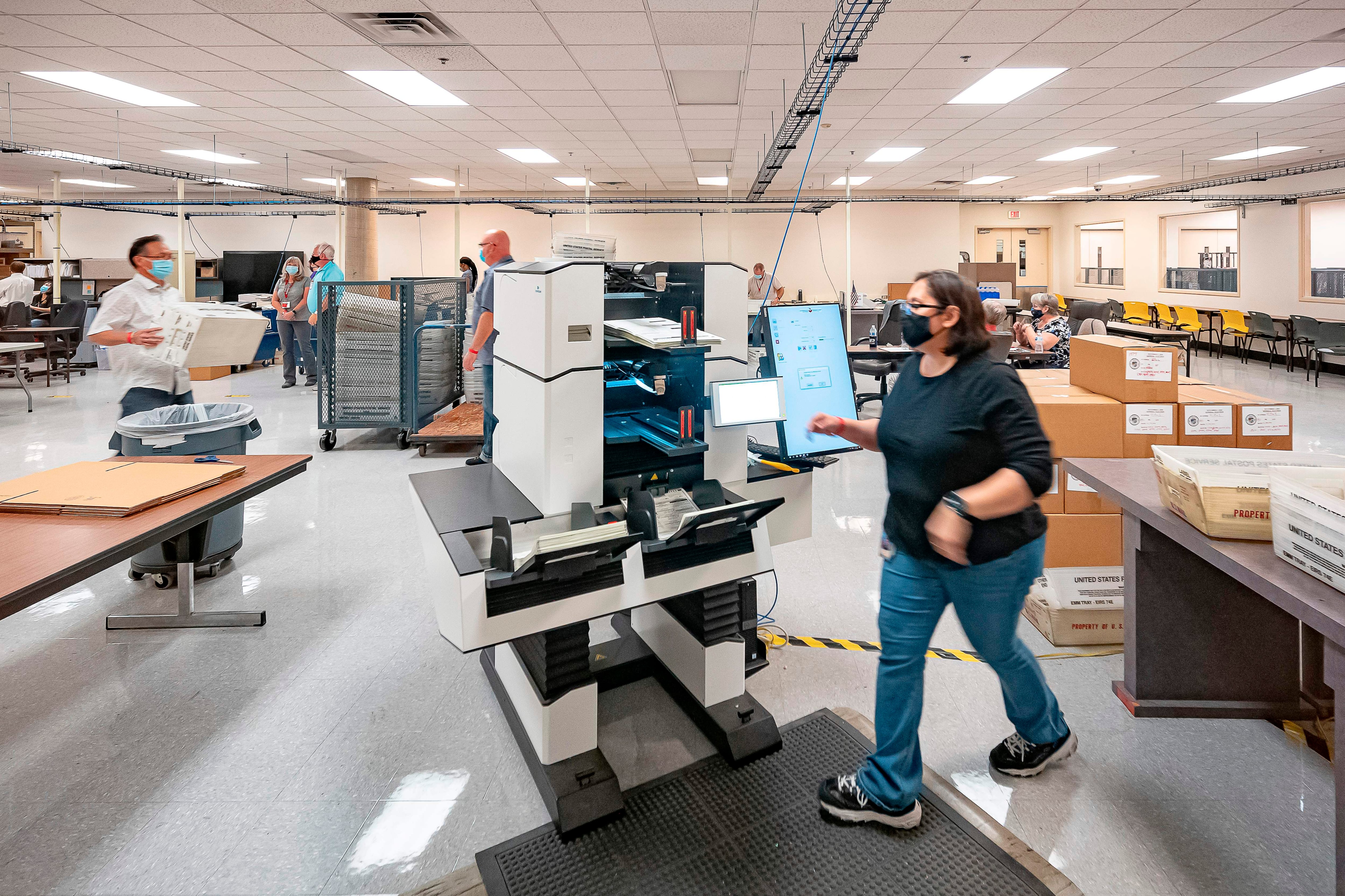 A worker approaches a machine in a large open room while other workers carry boxes and bins in the background.