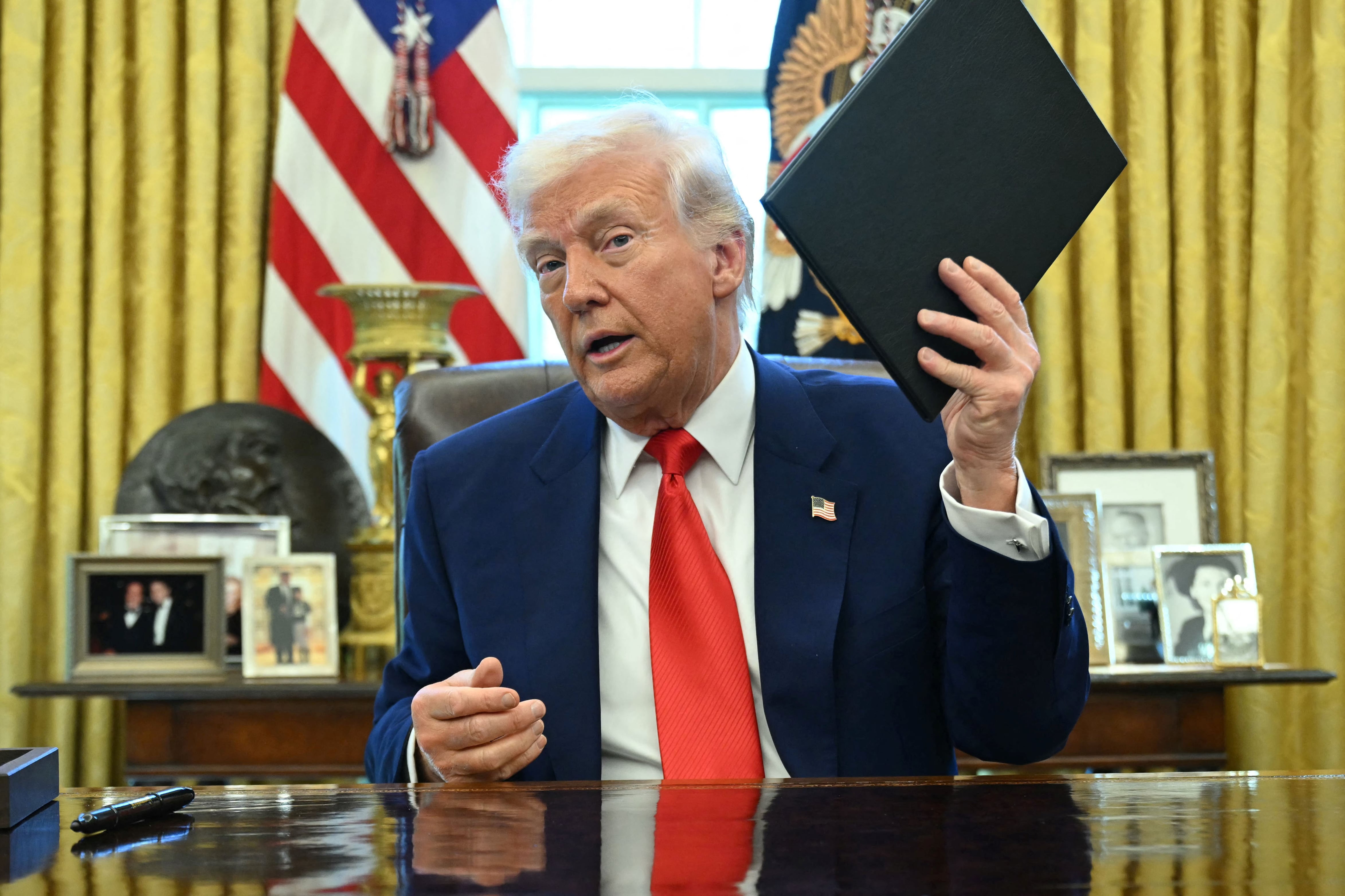 The president, a white man with white hair and wearing a blue suit and red tie looks at the camera while holding a booklet and sitting in the oval office.