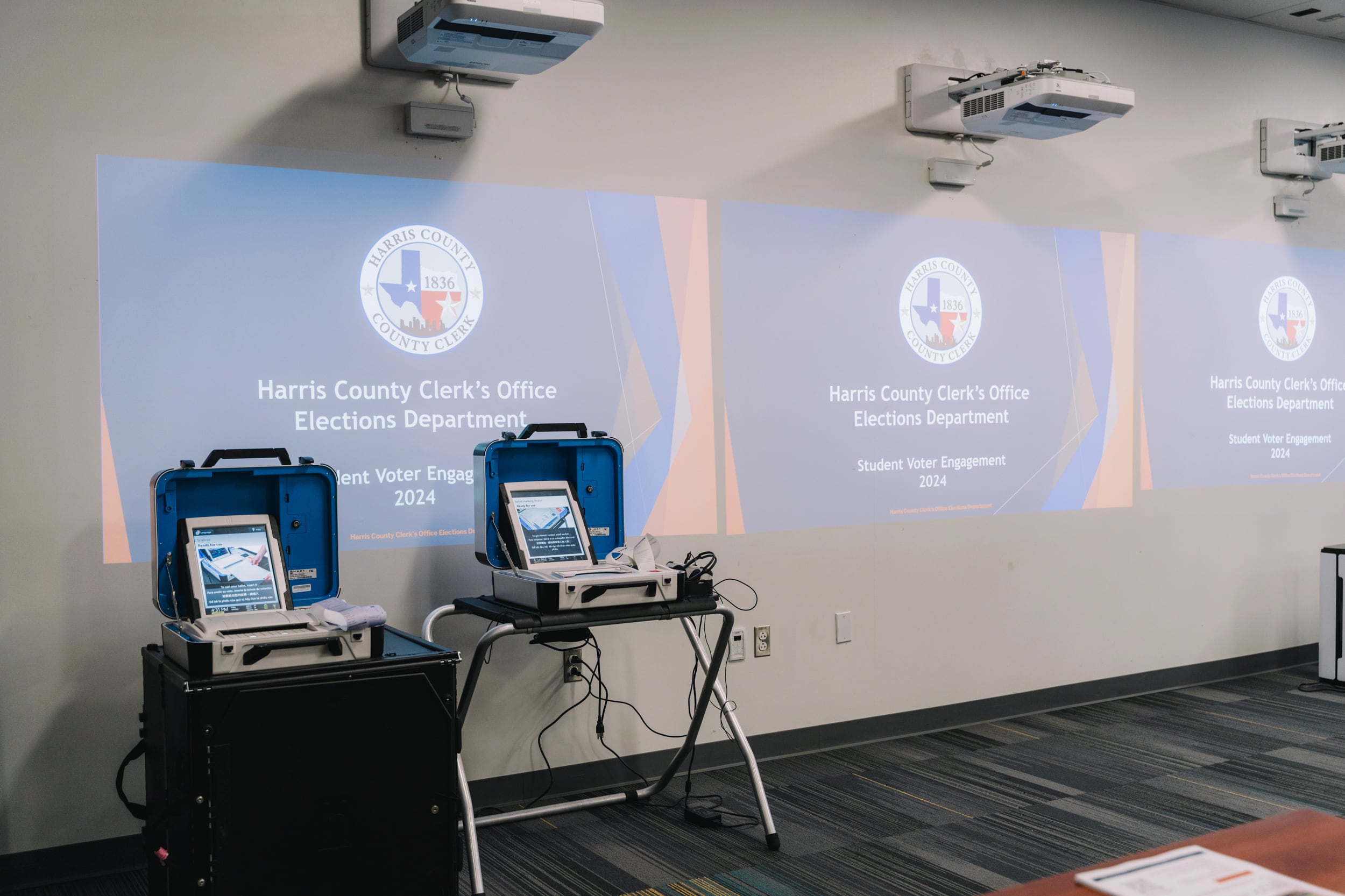 Three handing projectors project three identical images onto a plain wall with two voting machines standing up on the left of the image.