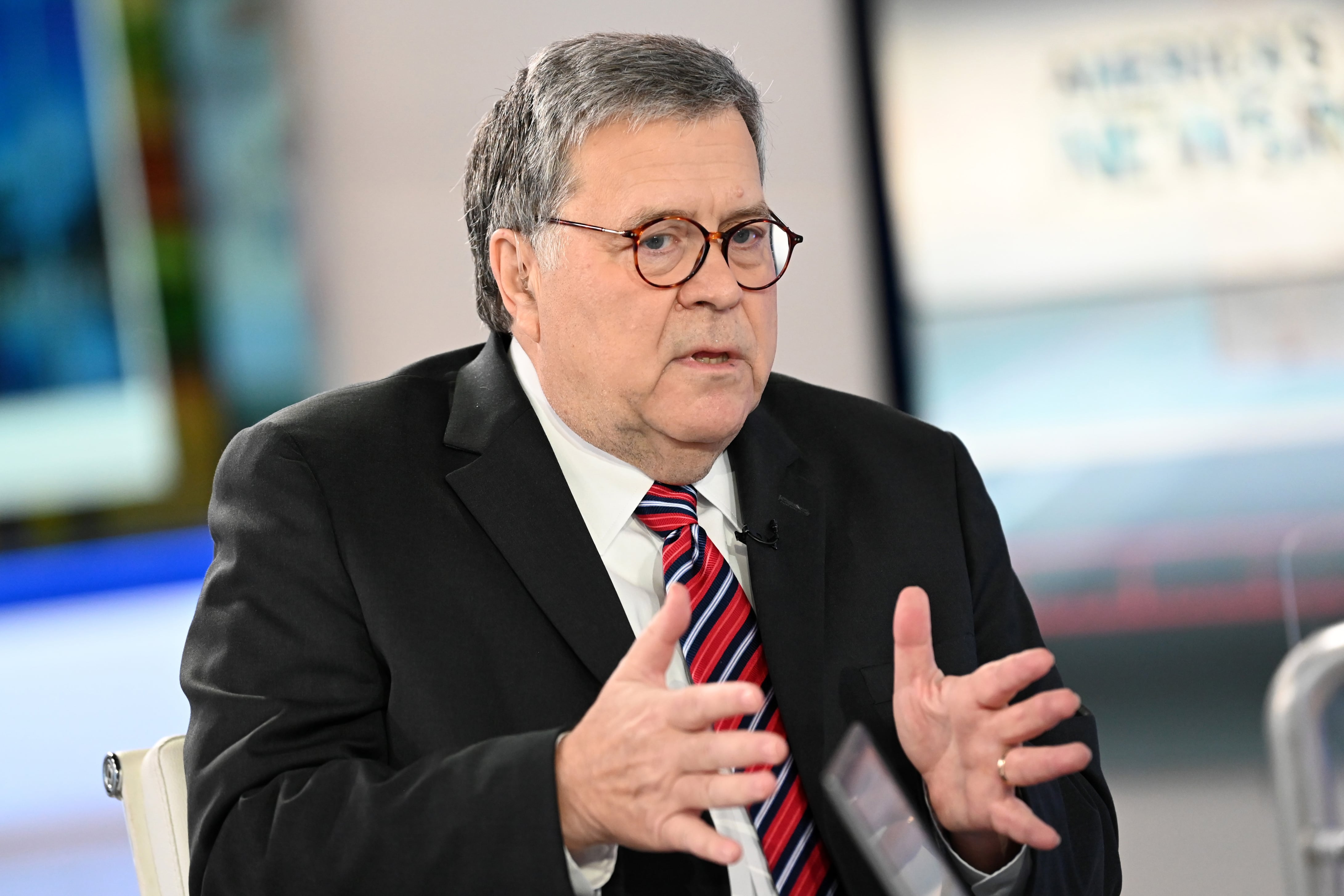 A man wearing glasses, a dark suit and a red tie motions with his hands while talking.