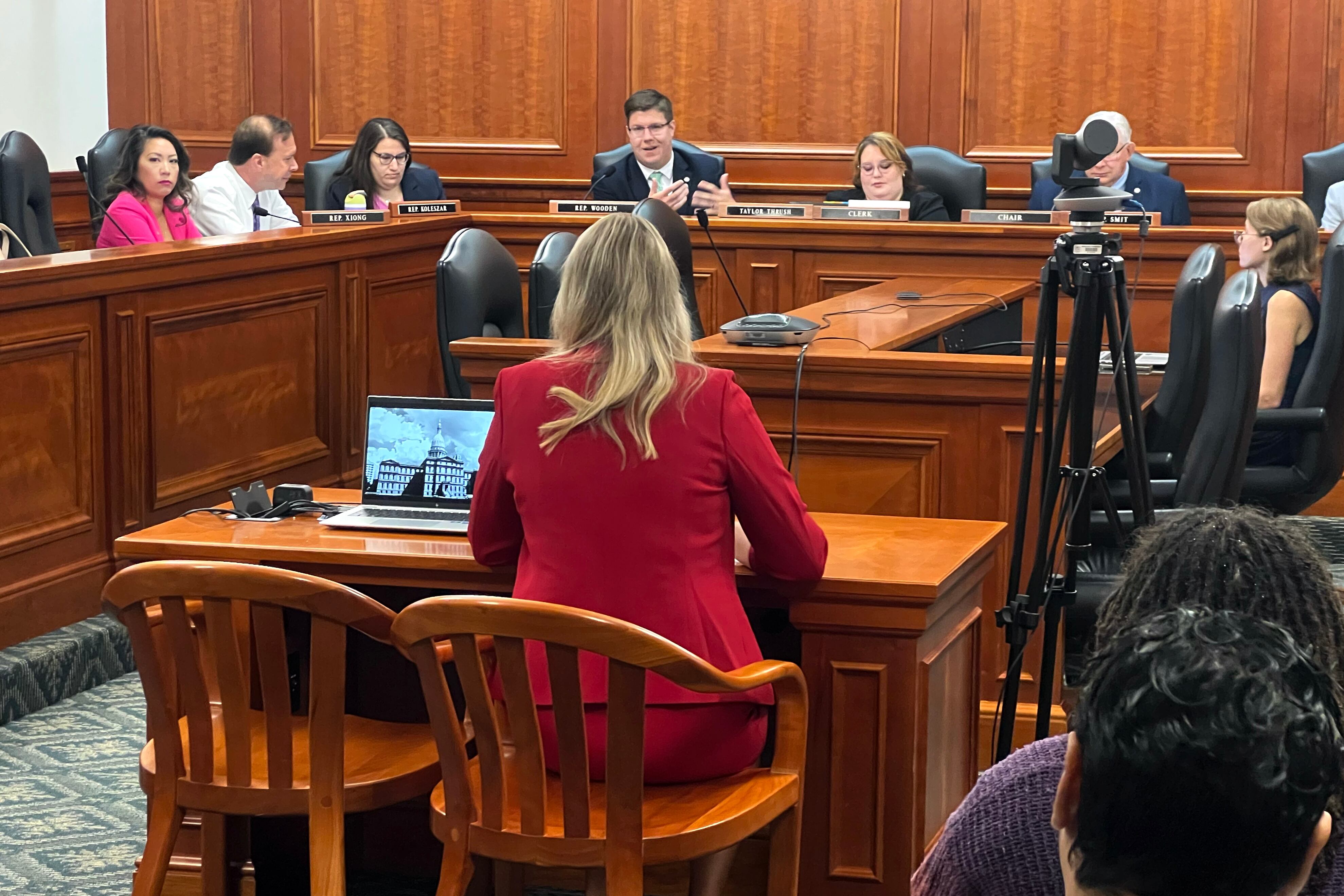 A photograph of a white woman in a red suit sitting at a wooden table facing a row of people in suits sitting at a large wooden table.