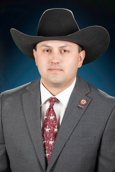 A man in a cowboy hat and wearing a grey suit poses for a photo with a dark green background.