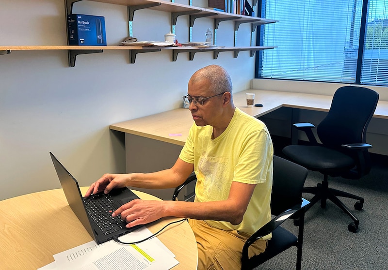 A man in a yellow t-shirt sits at a desk working on a laptop in an office.