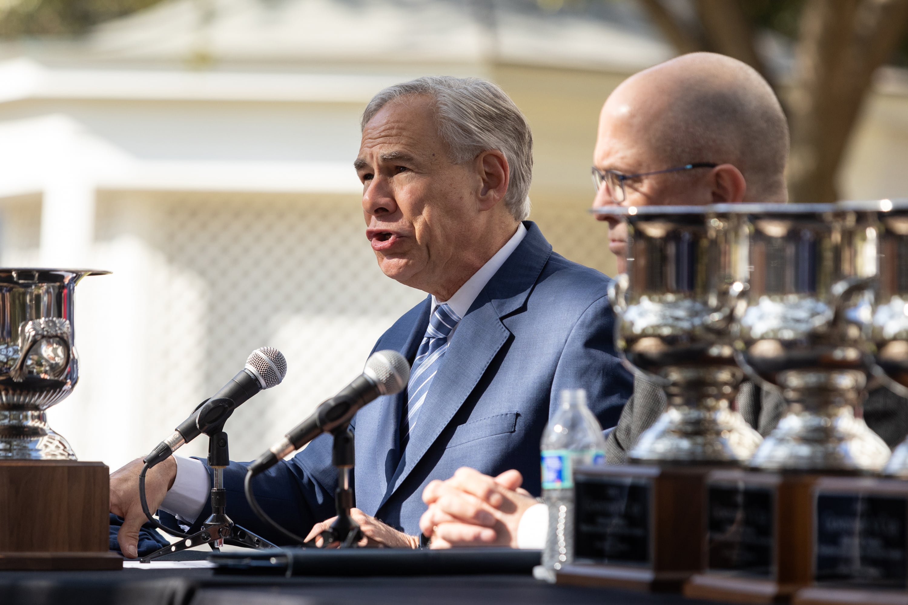 Two men sit at a table outside and speak from two microphones.