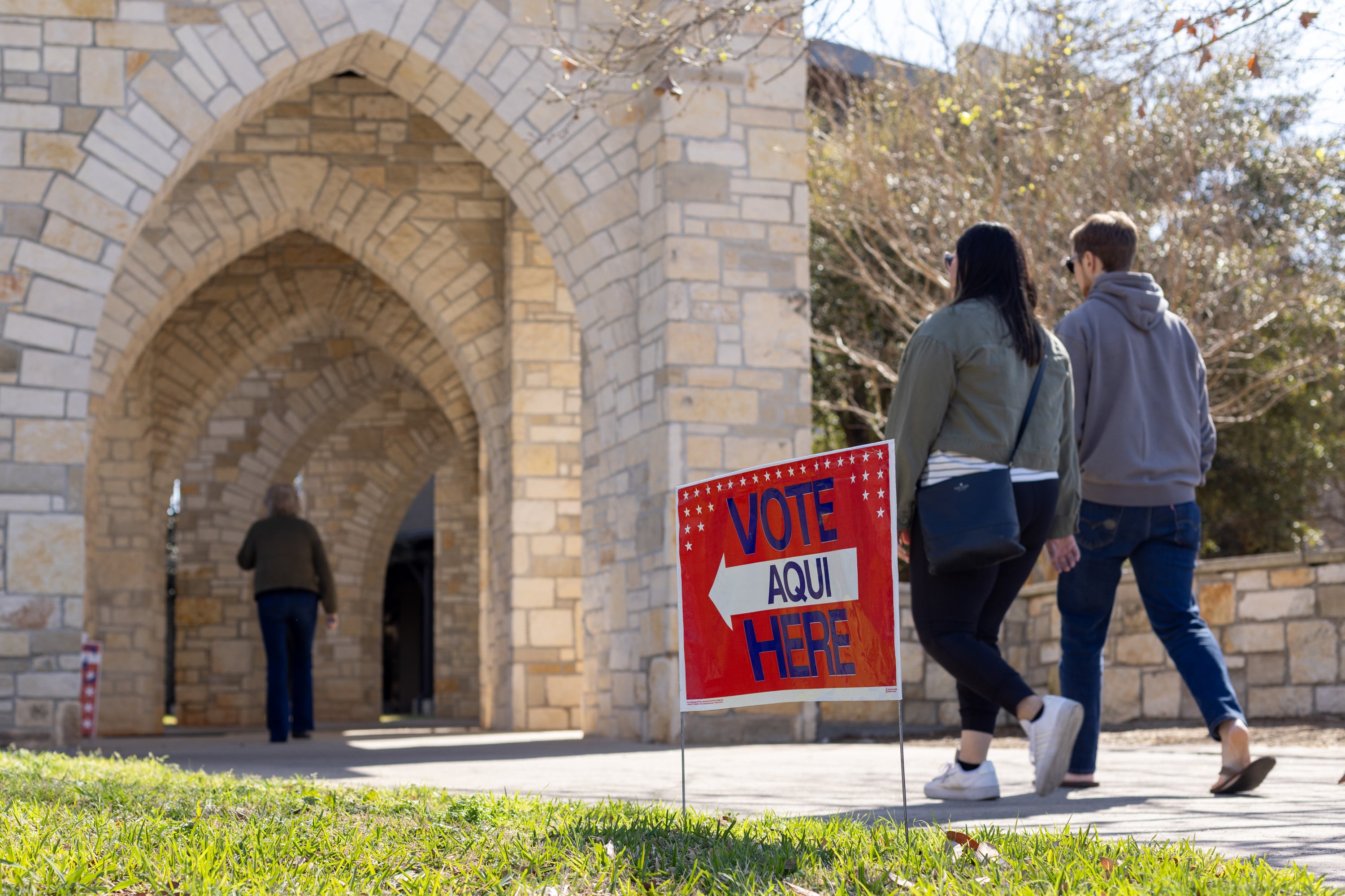 A photograph of two people walking next to a sign in the green grass next to the sidewalk that reads "Vote Here Aqui."
