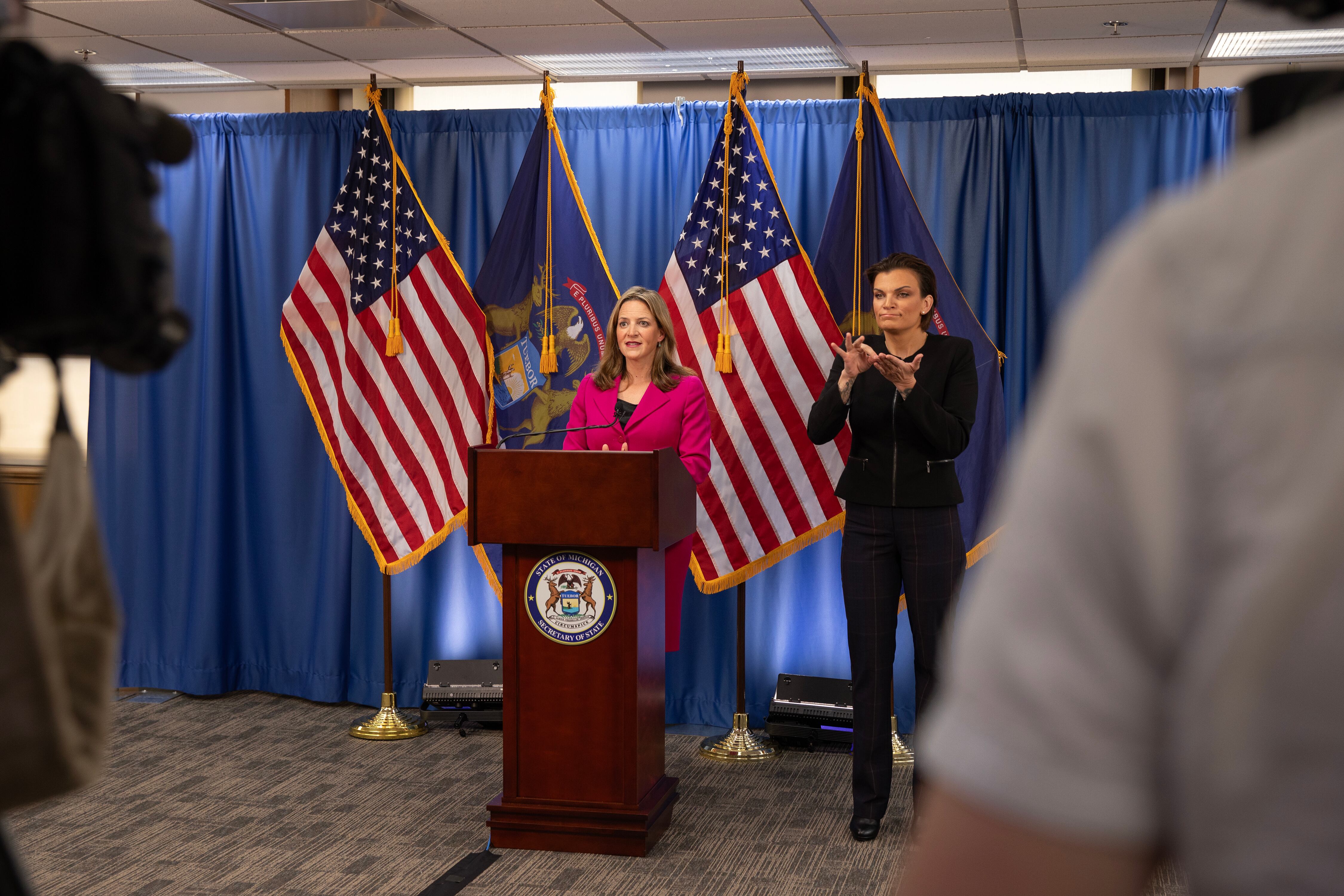 A woman with medium length blonde hair and wearing a bright pink suit speaks from behind a wooden podium and in front of a blue background with two American flags sandwiching a Michigan state flag and a woman with short dark hair and wearing a dark suit signs ASL on the right.