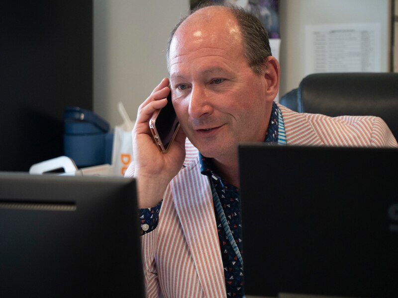 A man wearing a red and white striped suit sits at a desk behind computer screens and holds a phone to his ear.
